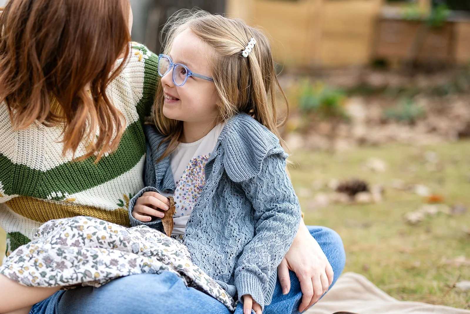 A woman and a young girl sitting on the grass in a park, smiling and engaging in conversation. The girl is wearing glasses and a gray knitted sweater, while the woman is wearing a striped sweater. The background shows blurred greenery and a wooden fe