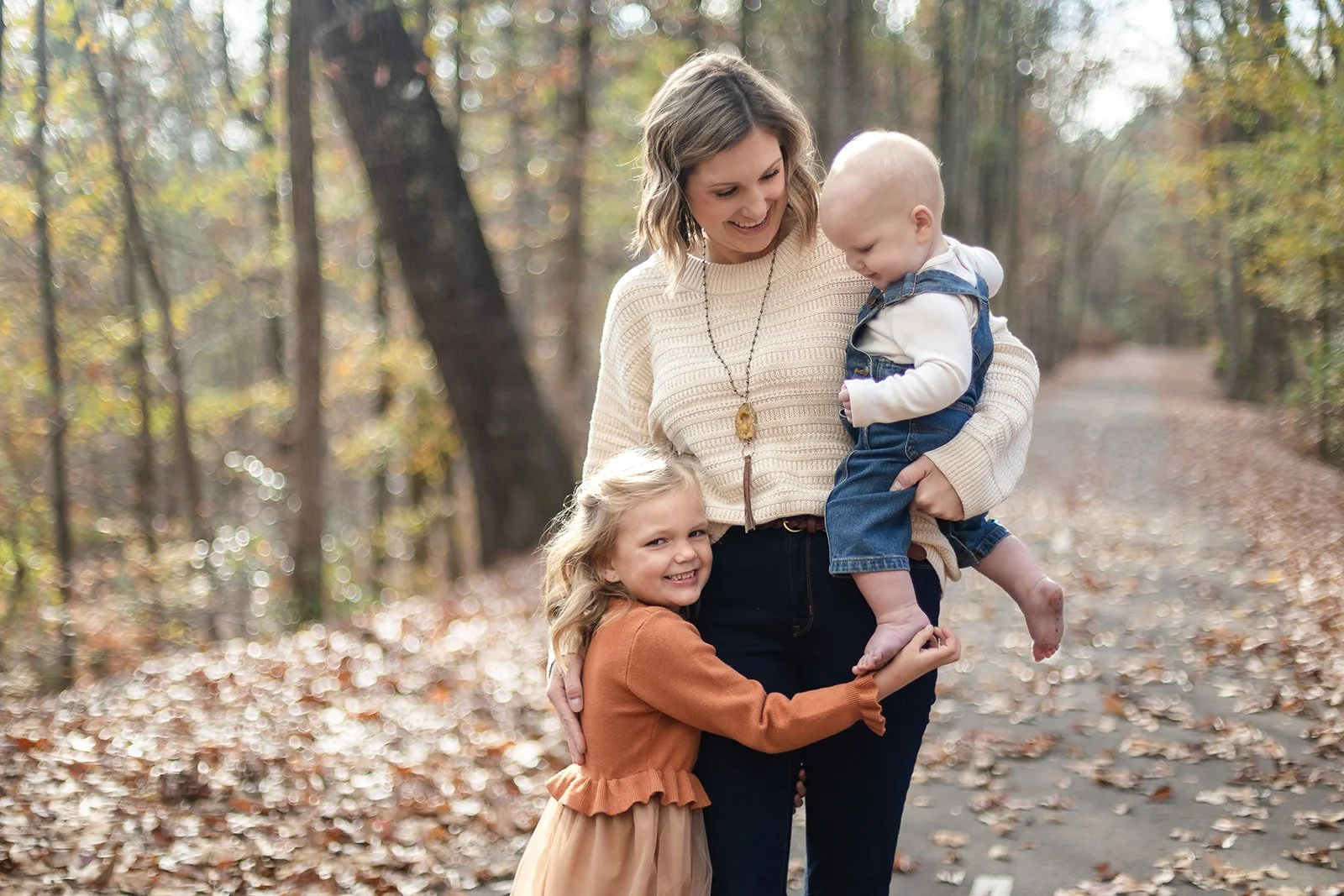 A woman and two children in a forest with fallen autumn leaves, smiling and playing together.