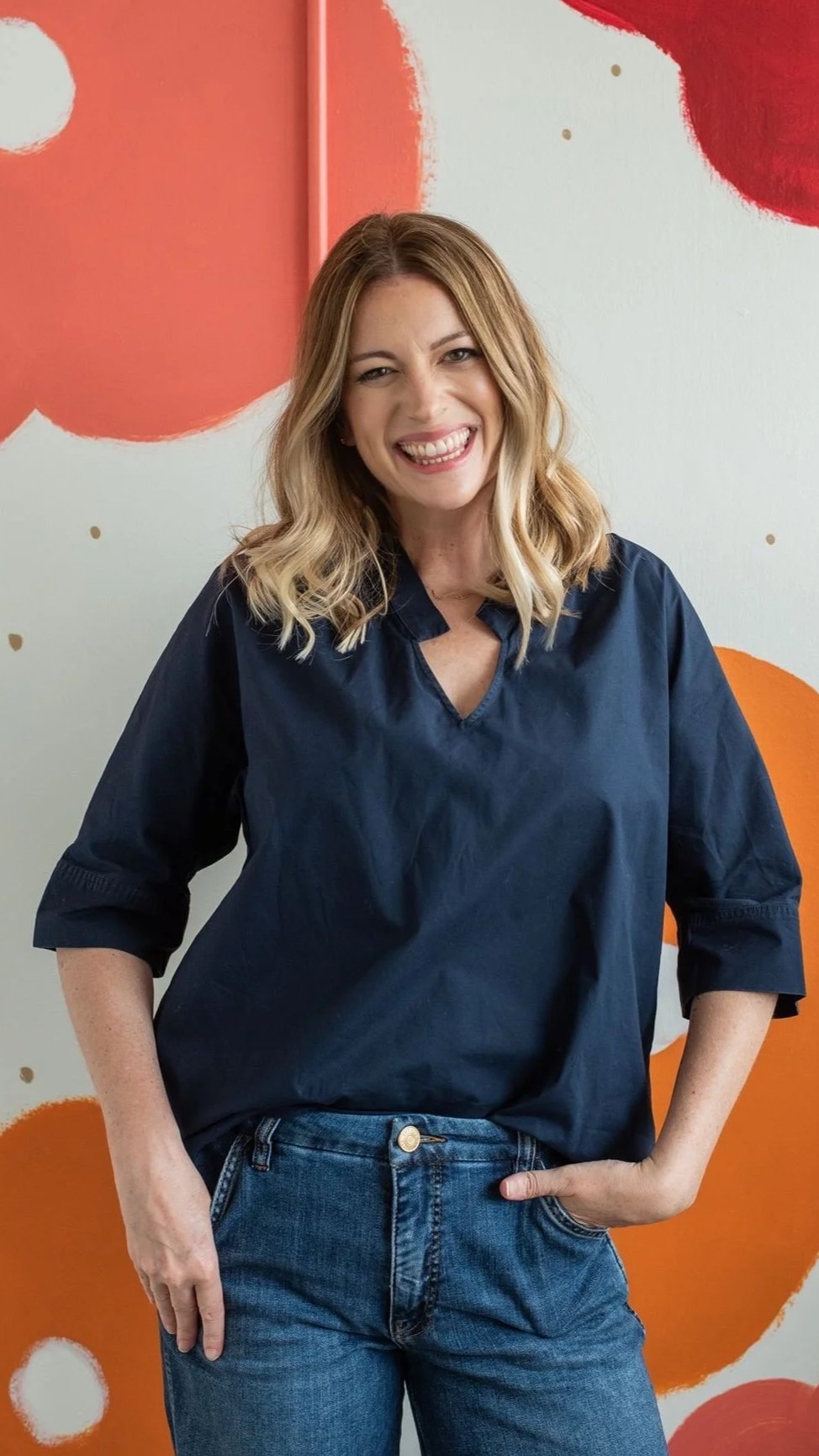 Sarah Watson with wavy blonde hair, wearing a dark blue shirt and blue jeans, smiling and posing with one hand on her hip in front of a colorful, abstract background.
