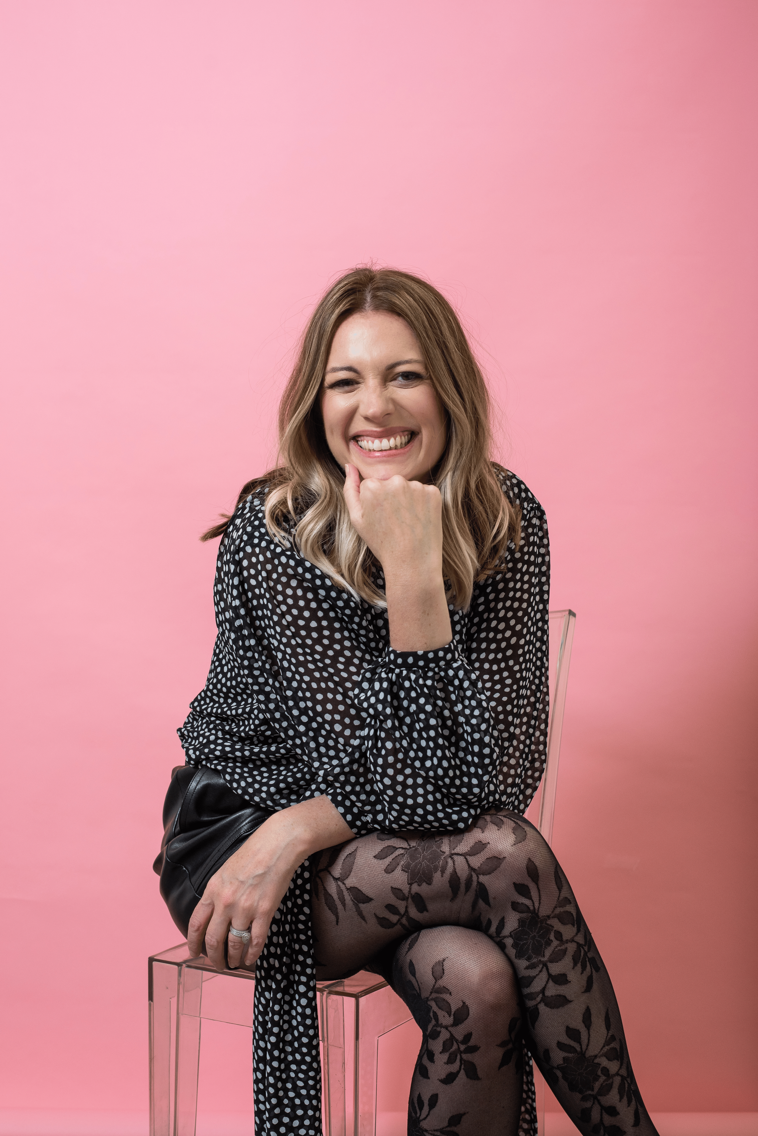 Sarah Watson sitting on a transparent chair against a pink background, smiling, wearing a black dress with white polka dots, floral patterned thigh-high stockings, and a ring on her left hand.