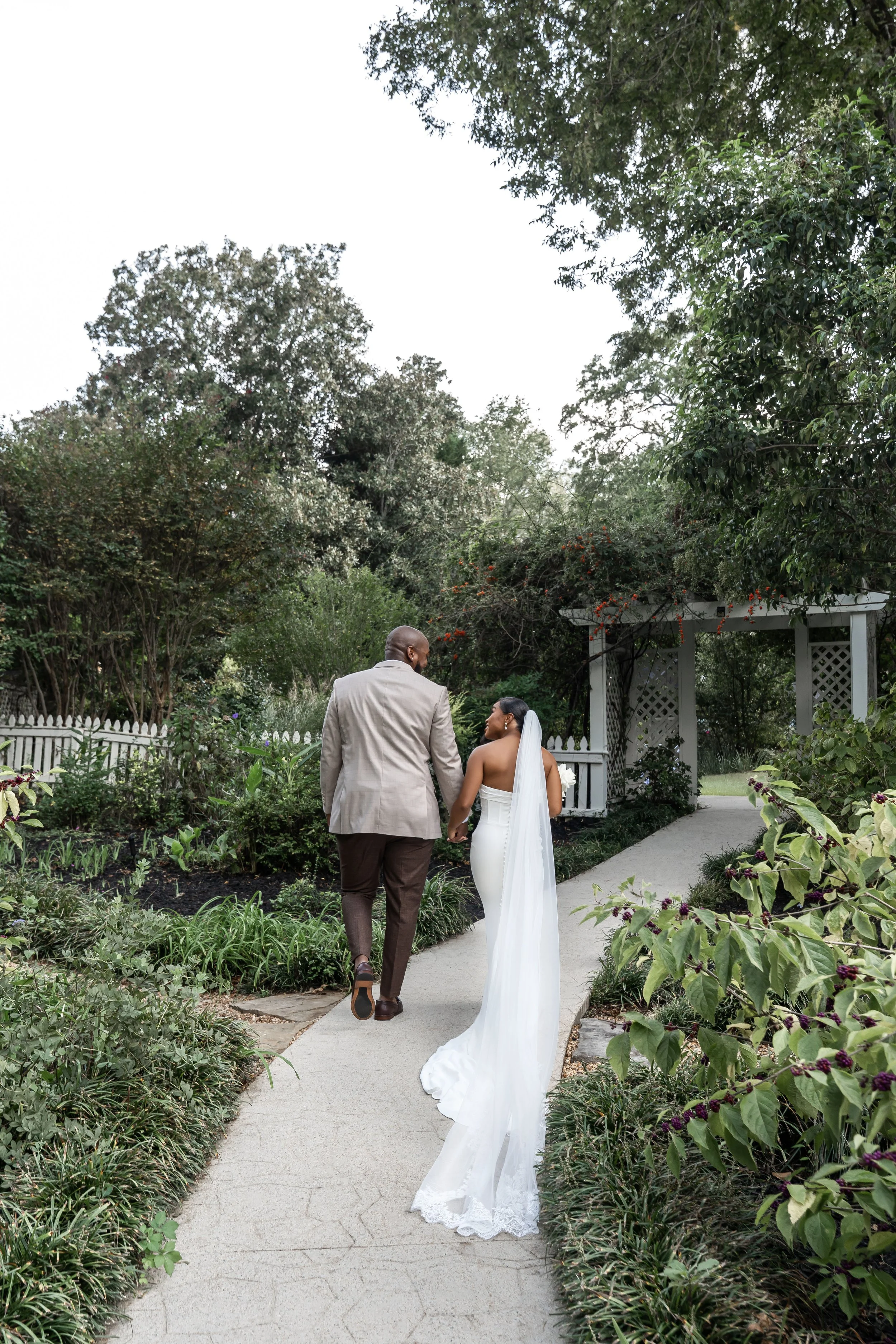 A bride and groom walking hand in hand on a garden pathway during their wedding day.