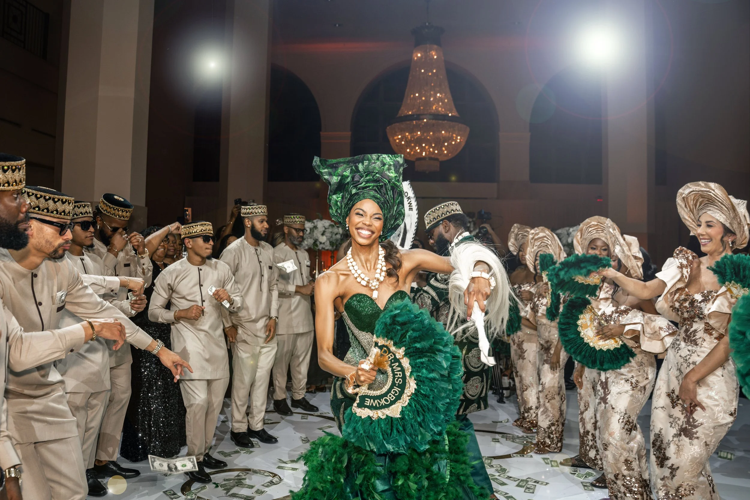 An African bride in a green dress and headwrap dances during her wedding reception, surrounded by men and women in traditional attire with headwraps, in a decorated event hall with a chandelier in the background, with dollar bills on the floor.