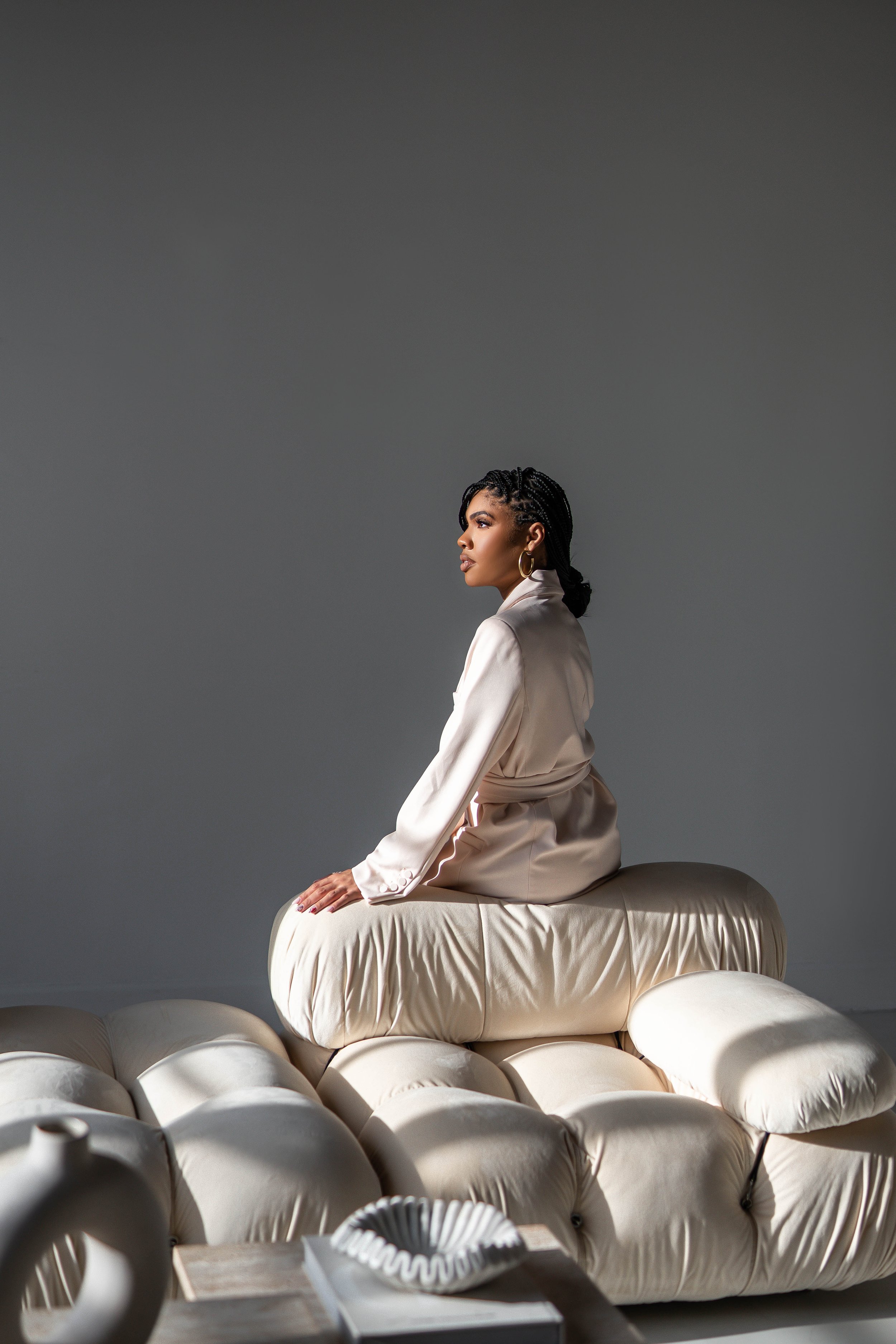 A woman in a beige satin robe sitting on rounded, beige tufted cushions, with a plain gray background.