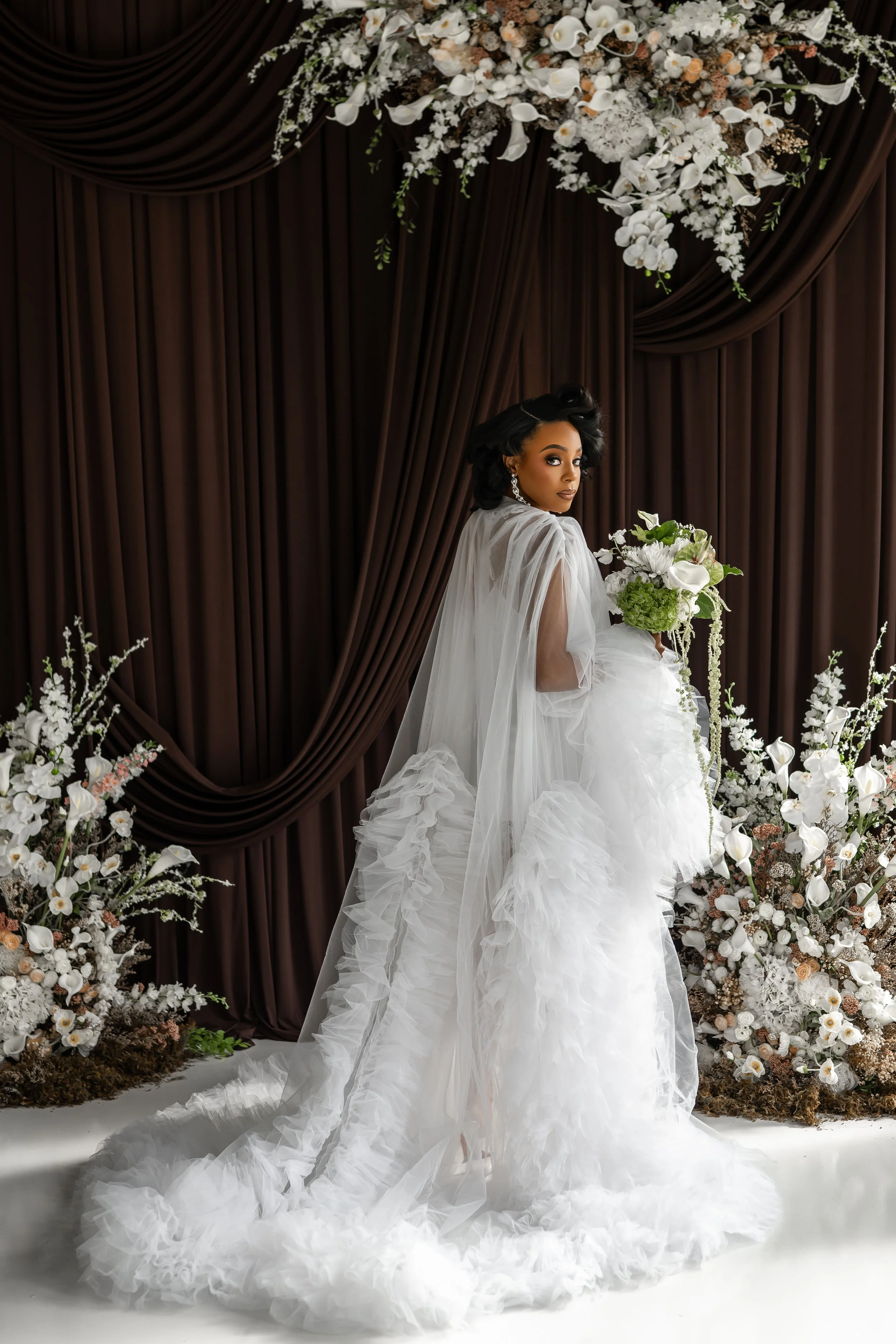 A woman in a white wedding dress holding a bouquet of white and green flowers, standing in front of a backdrop with dark brown drapes and white floral arrangements.
