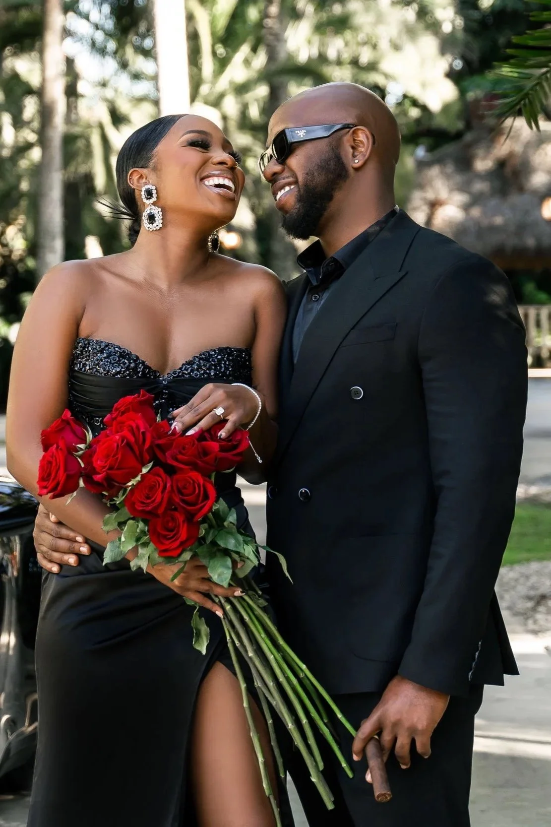 A couple dressed in formal attire, smiling and sharing a moment outdoors; the woman holds a bouquet of red roses and is wearing a strapless black gown with a high slit, while the man wears a black suit and sunglasses.