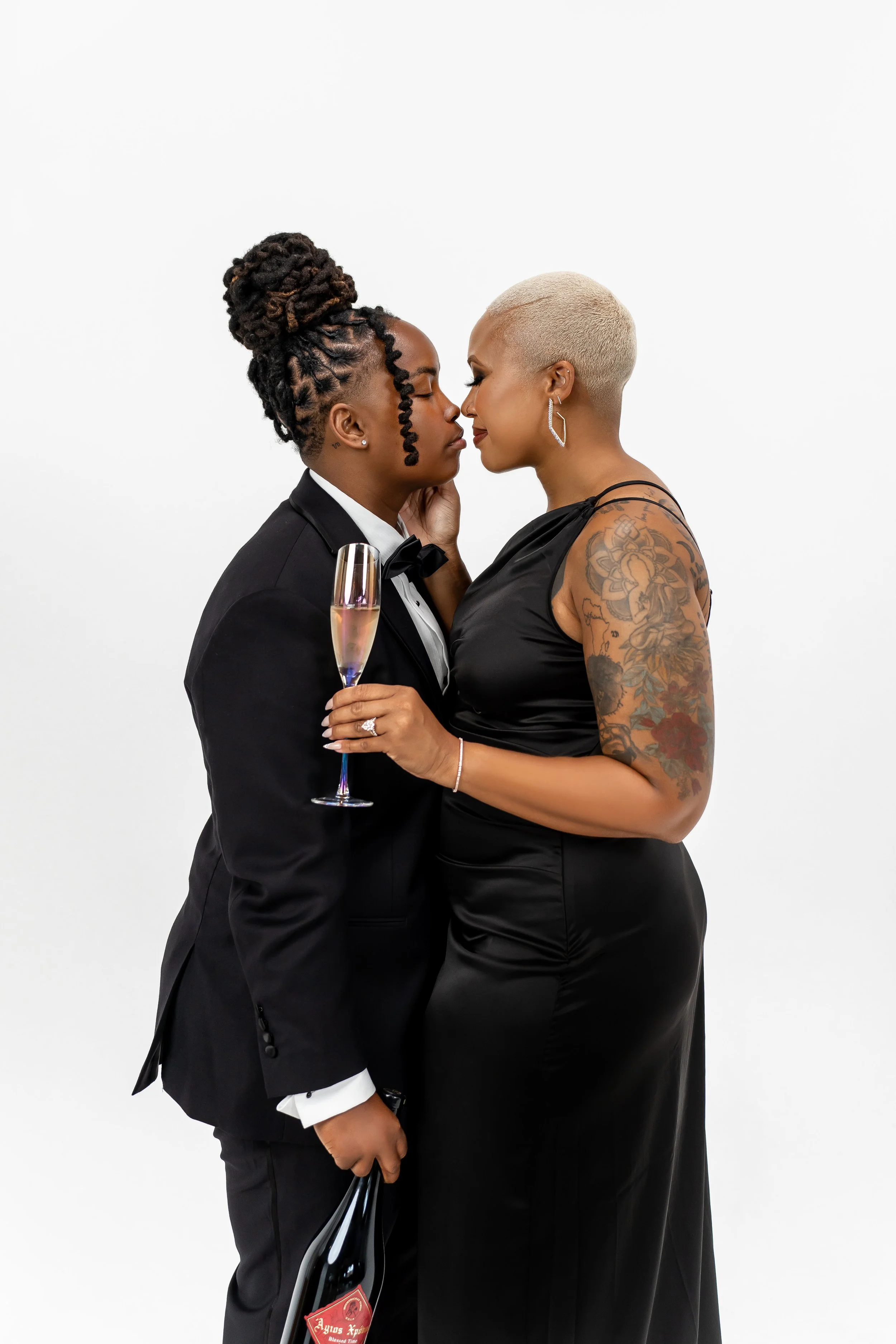 Two women embracing, one in a tuxedo holding a glass of champagne and a bottle, the other in a black dress with tattoos on her arm, against a plain white background.