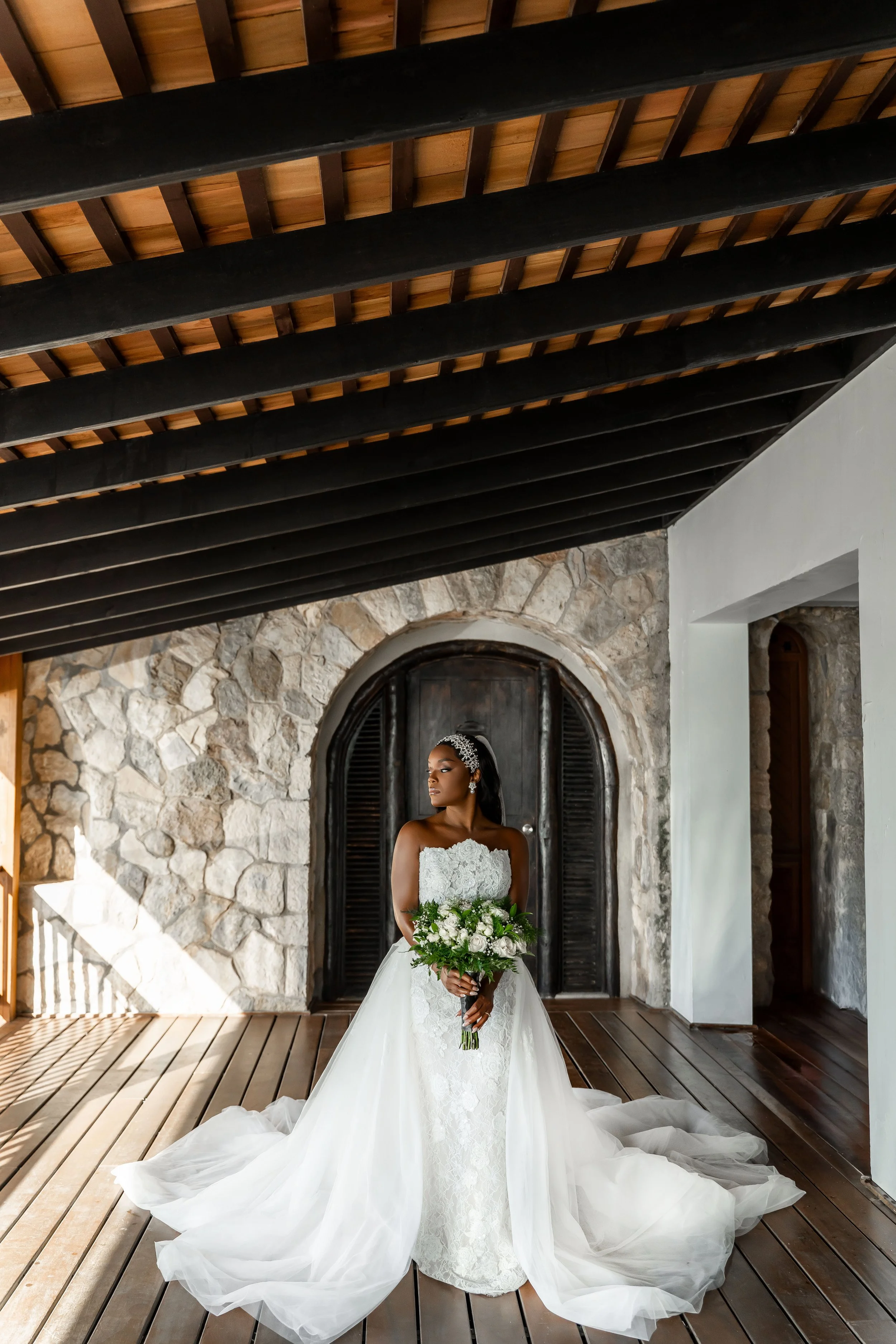 Bride in a white wedding dress holding a bouquet of white flowers, standing on a wooden floor in front of a stone wall with a black wooden door, sunlight streaming in from the side. Pattoo Castle Wedding in Jamaica. 