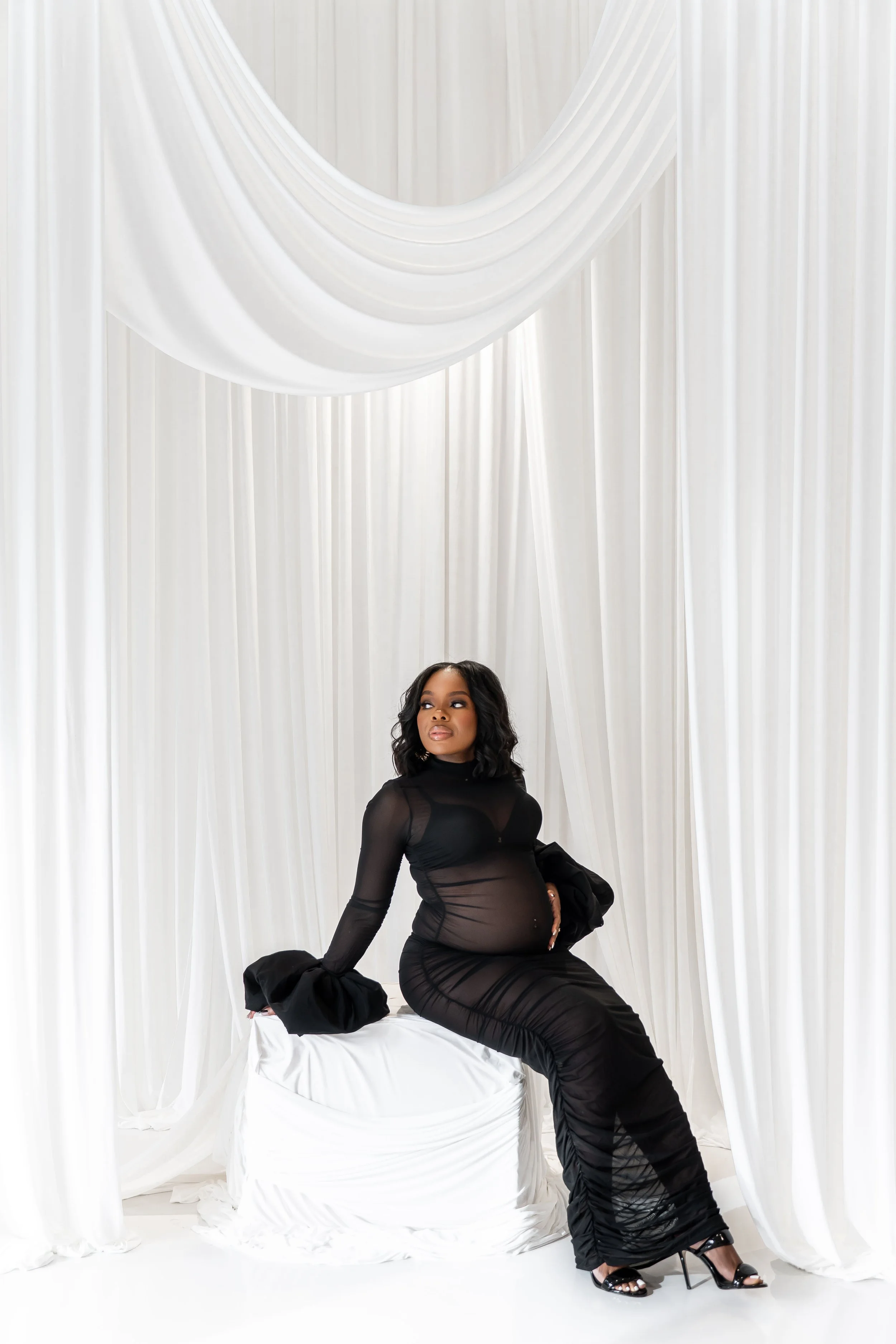 A woman in a sheer black dress and high heels sitting on a white covered surface against a backdrop of white draped curtains.