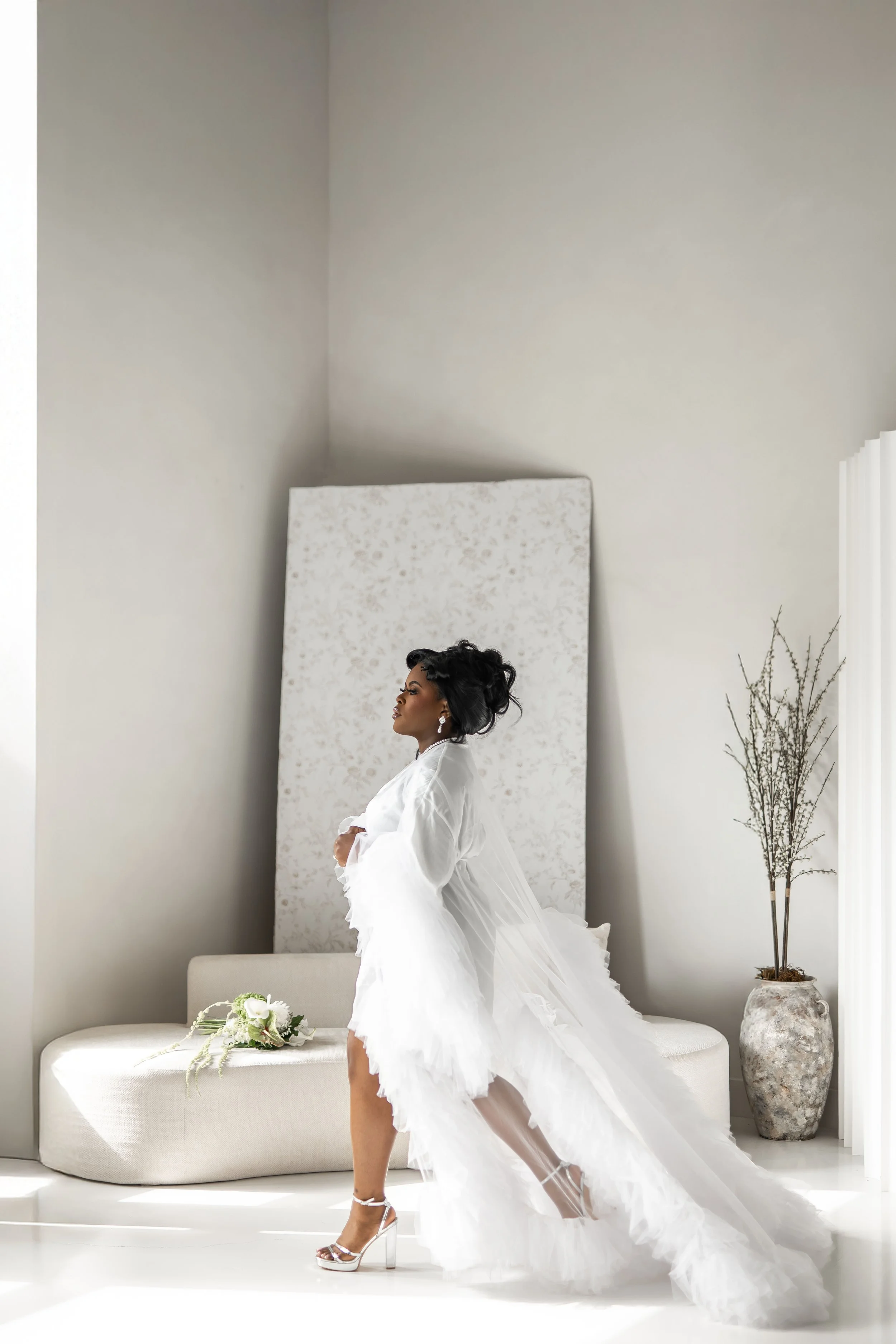 A bride wearing a white wedding dress and high heels standing in a bright, minimalistic room with a floral bouquet on a beige couch and a large potted plant in a textured vase.