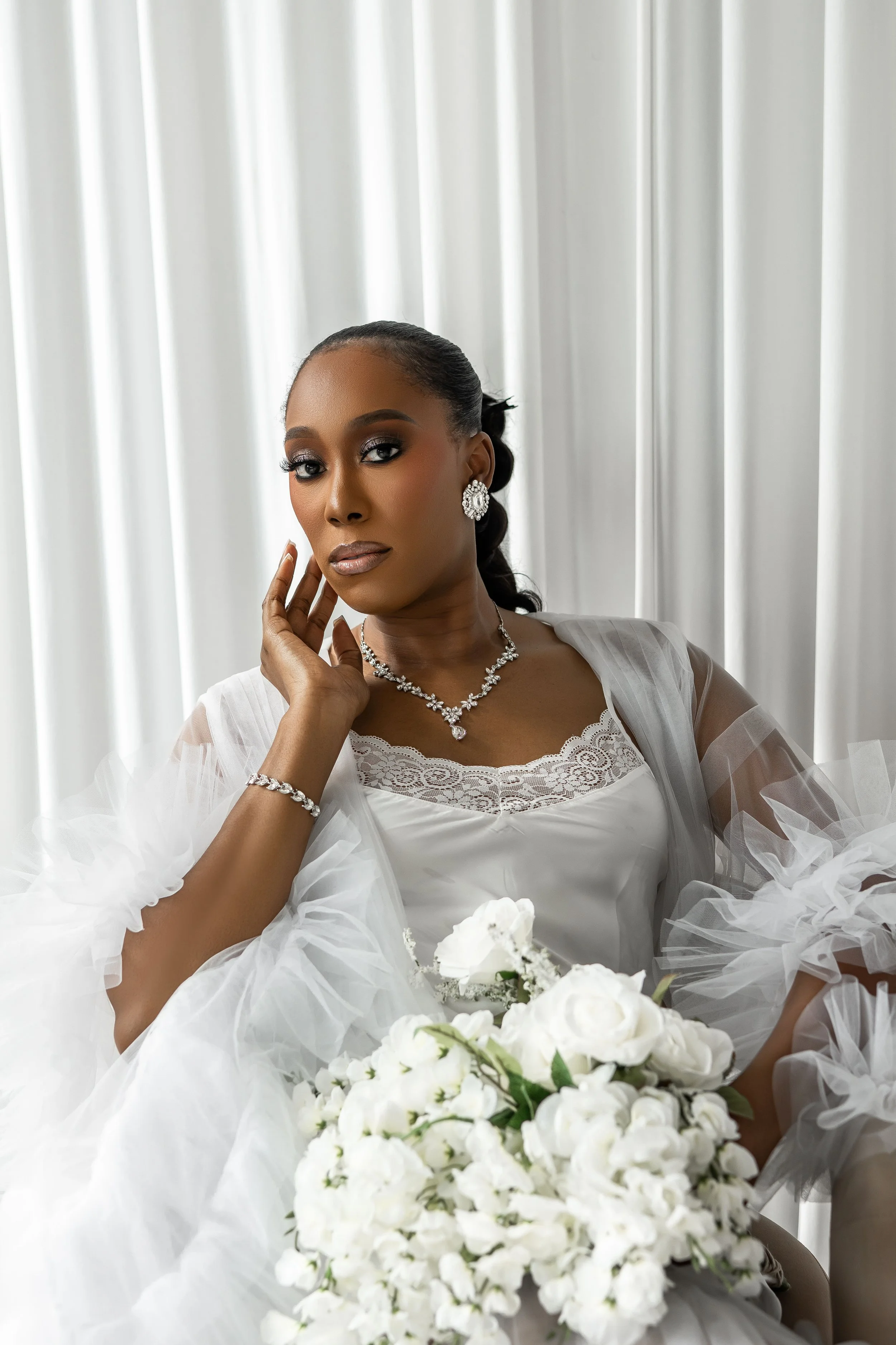 A woman dressed in white with jewelry, holding a bouquet of white flowers, with a contemplative expression, in front of a white curtain.
