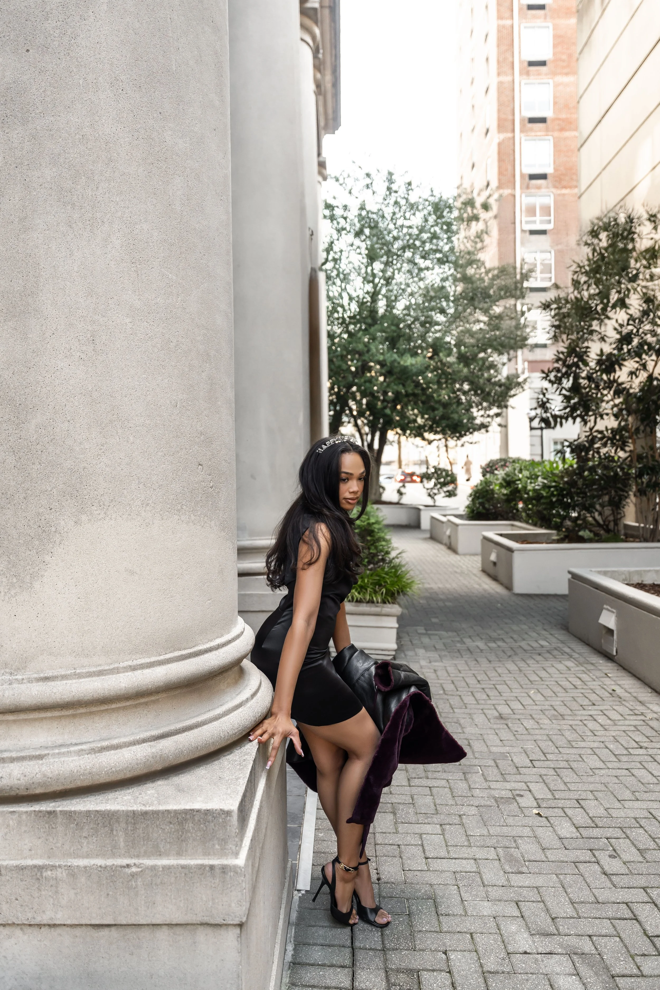 Woman in black dress and high heels standing next to a large stone column on a city sidewalk with trees and buildings in the background.