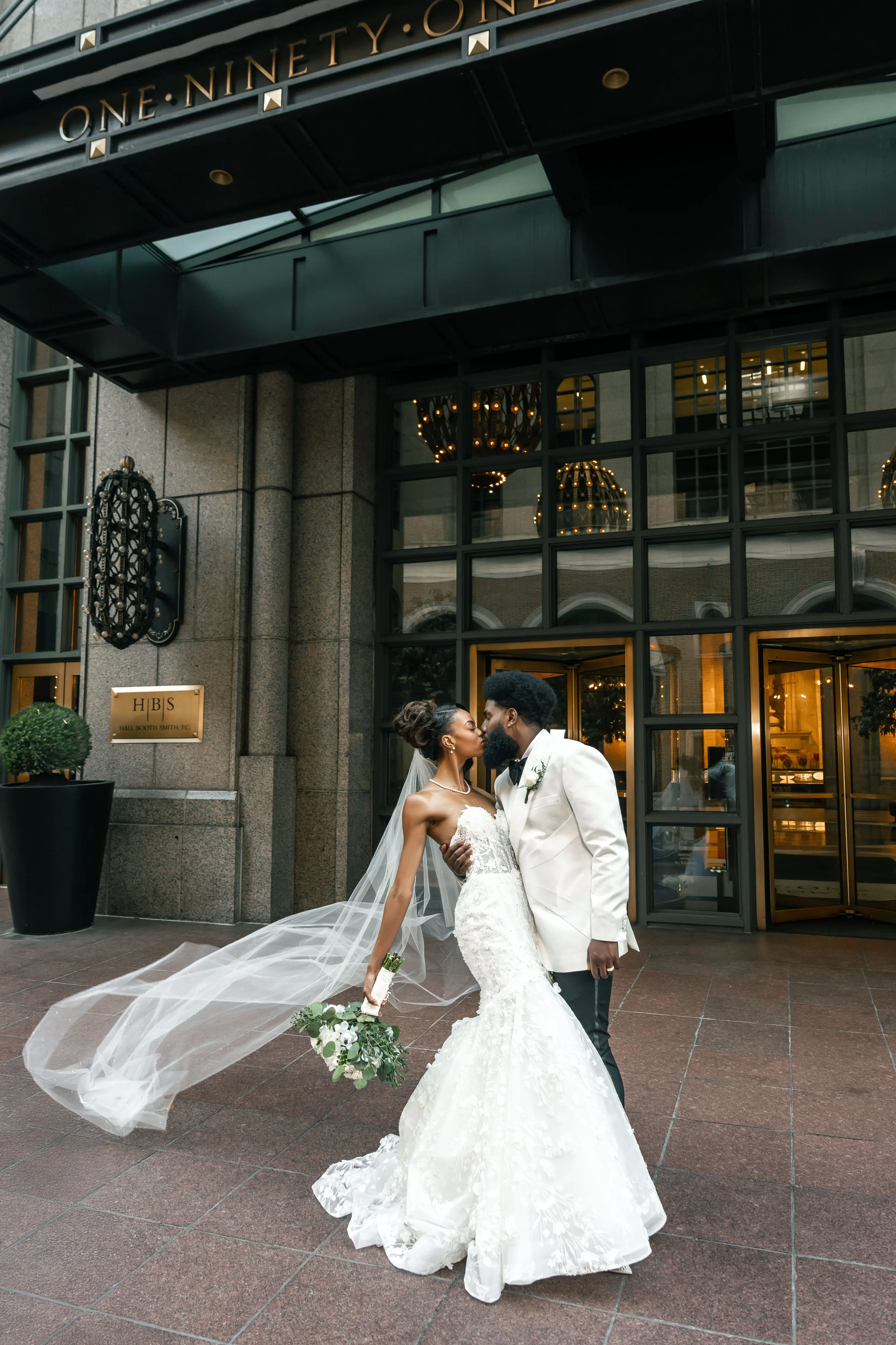 Bride and groom sharing a kiss after their New York City Wedding. The bride is in a white wedding gown holding a bouquet, and the groom is in a white tuxedo jacket. The building has large glass doors and windows with warm lighting inside.