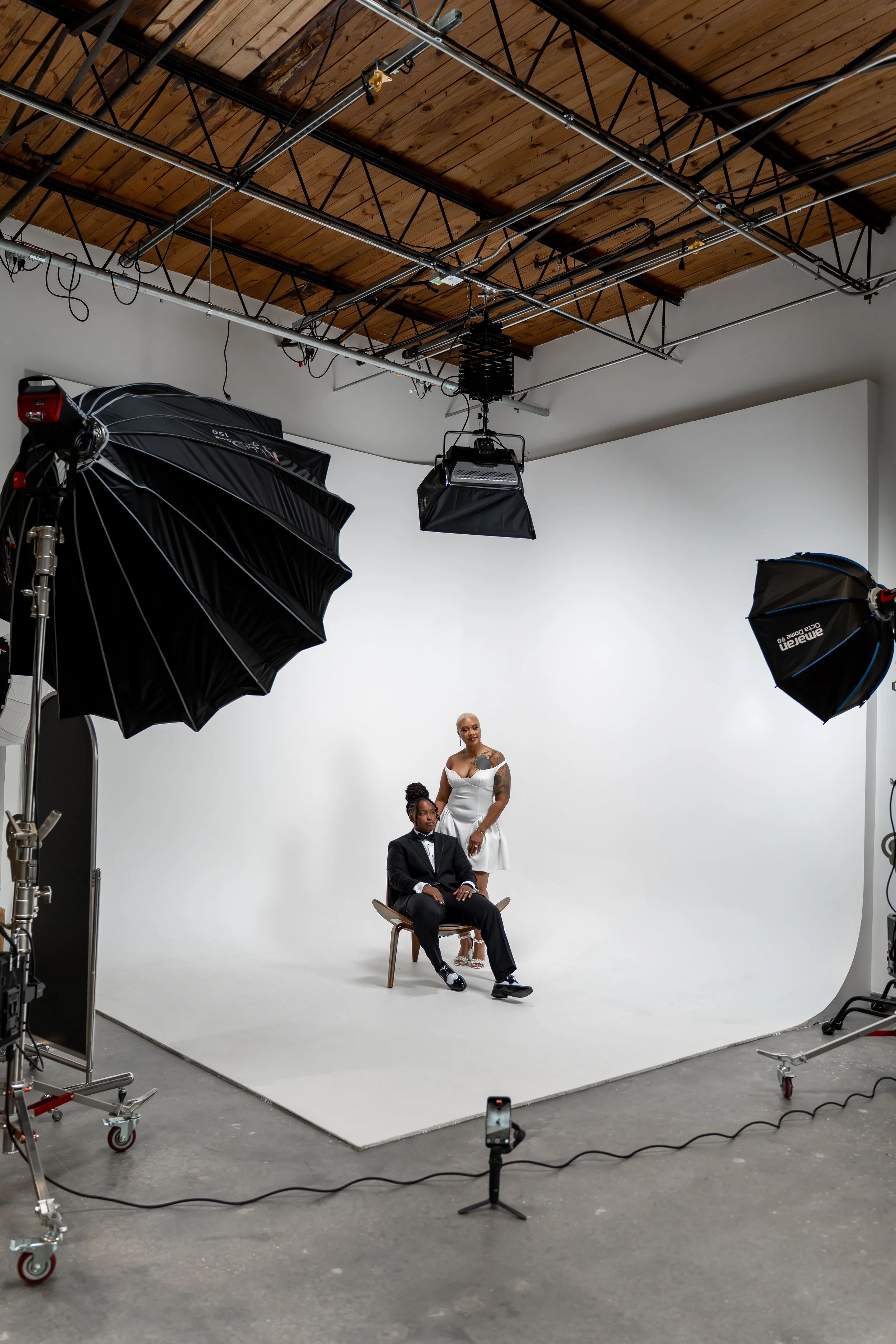 A photoshoot setup with two models in formal attire, one seated and the other standing, in a white studio with professional lighting and equipment.