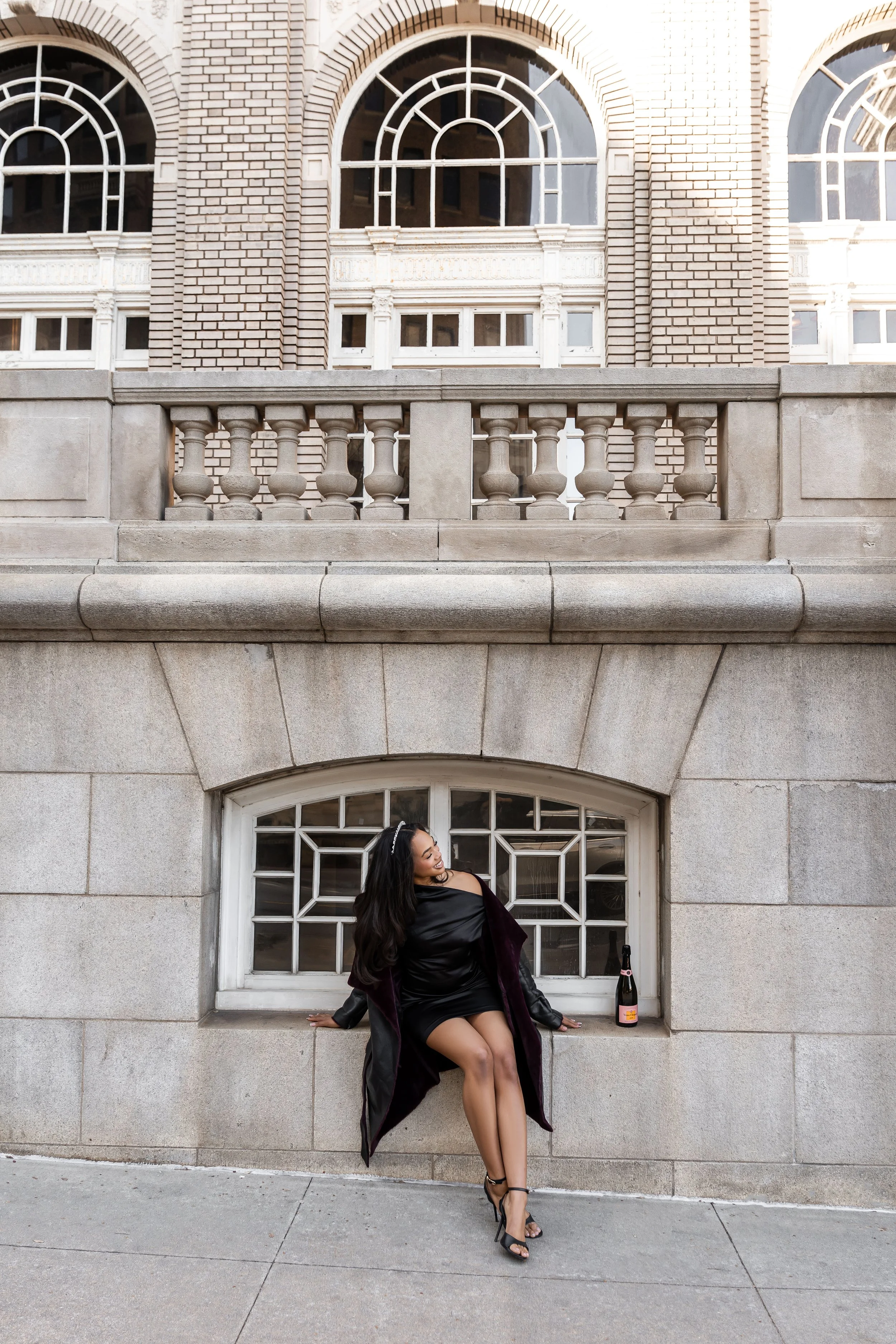 A woman in a black dress and high heels sitting on a ledge in front of a large stone building with arched windows, a bottle of champagne beside her, looking to the side and smiling.