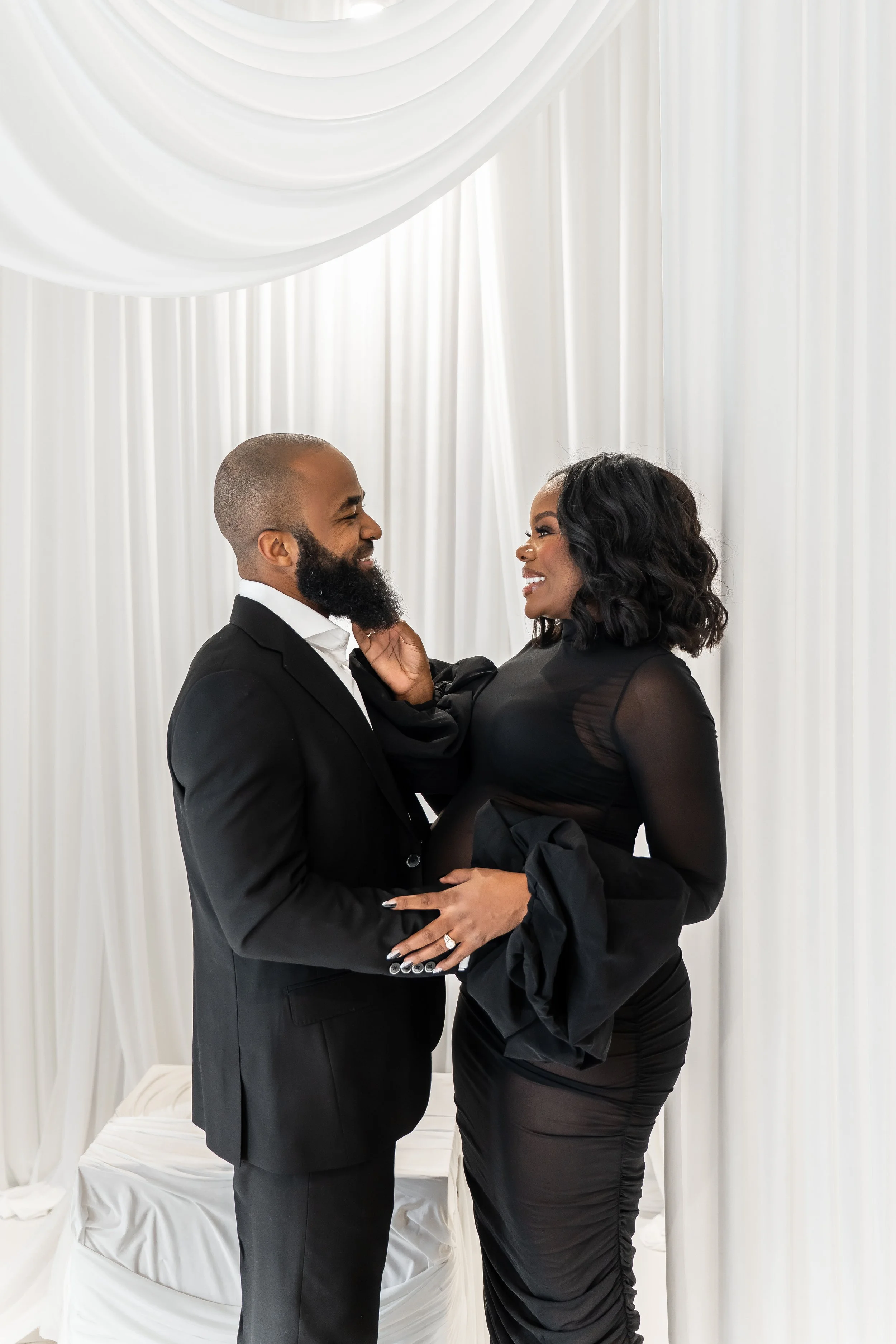 A couple dressed in formal attire sharing a joyful moment indoors, with white draped curtains in the background.