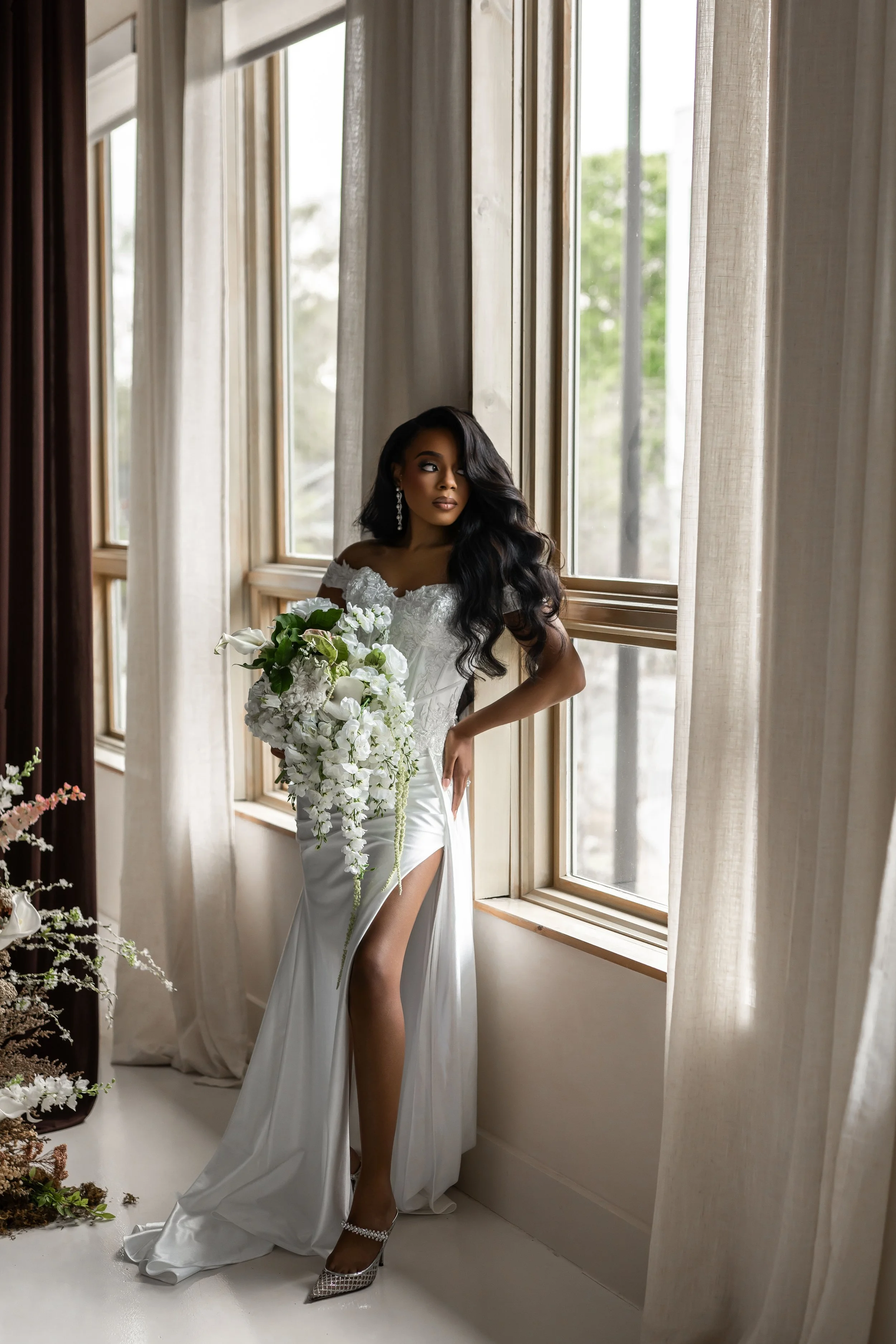 A bride in a white wedding dress with a slit, holding a large bouquet of white flowers, standing by a window with beige curtains inside a well-lit room.