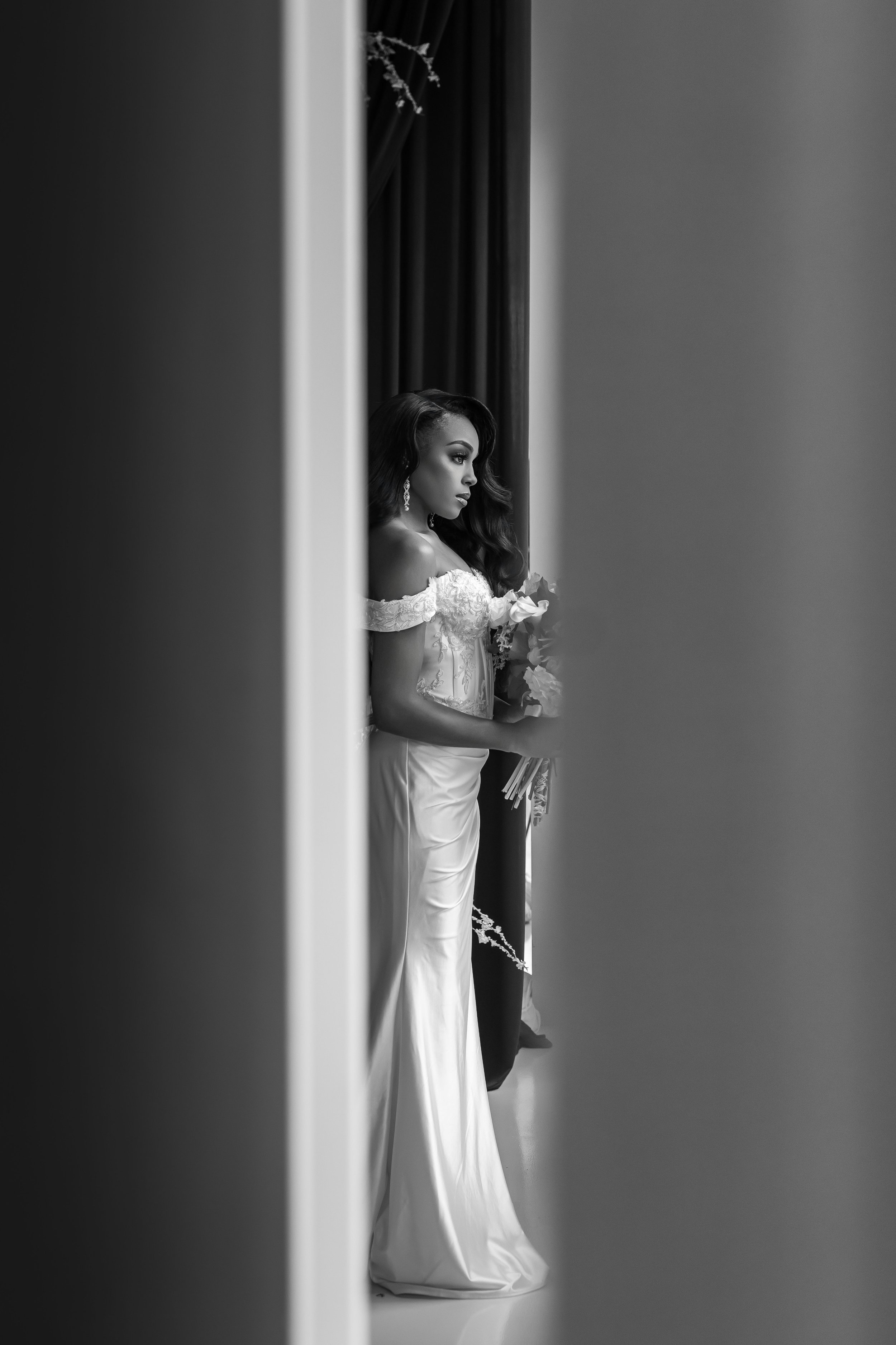 Black and white photo of a bride in an elegant dress holding a bouquet, viewed through a partially open door with dark curtains behind her.