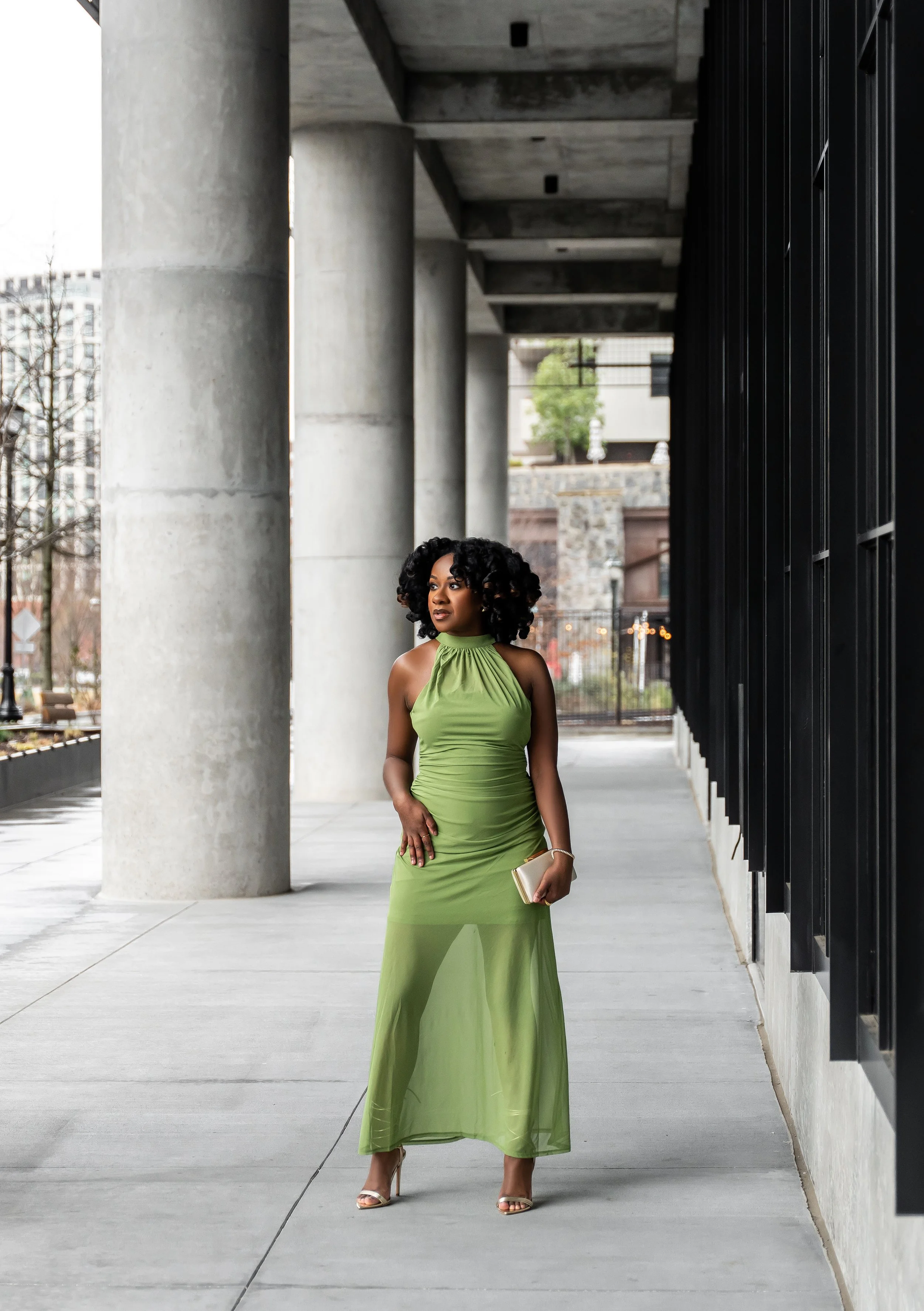 A woman in a long, green halter dress standing outside on a sidewalk under a concrete shelter, holding a gold clutch and wearing high heels.