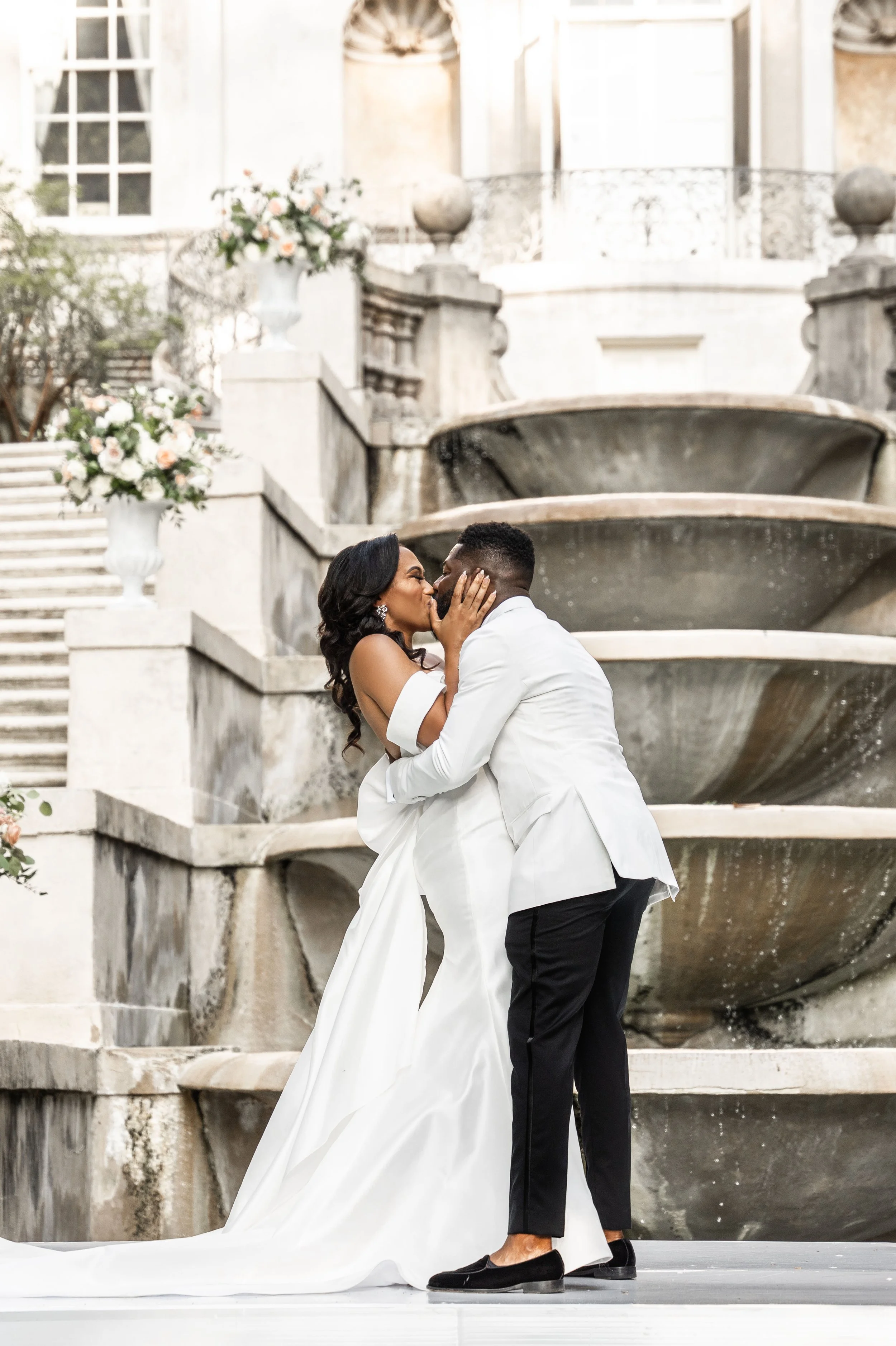 Couple in wedding attire kissing in front of a stone fountain and staircase with floral decorations.