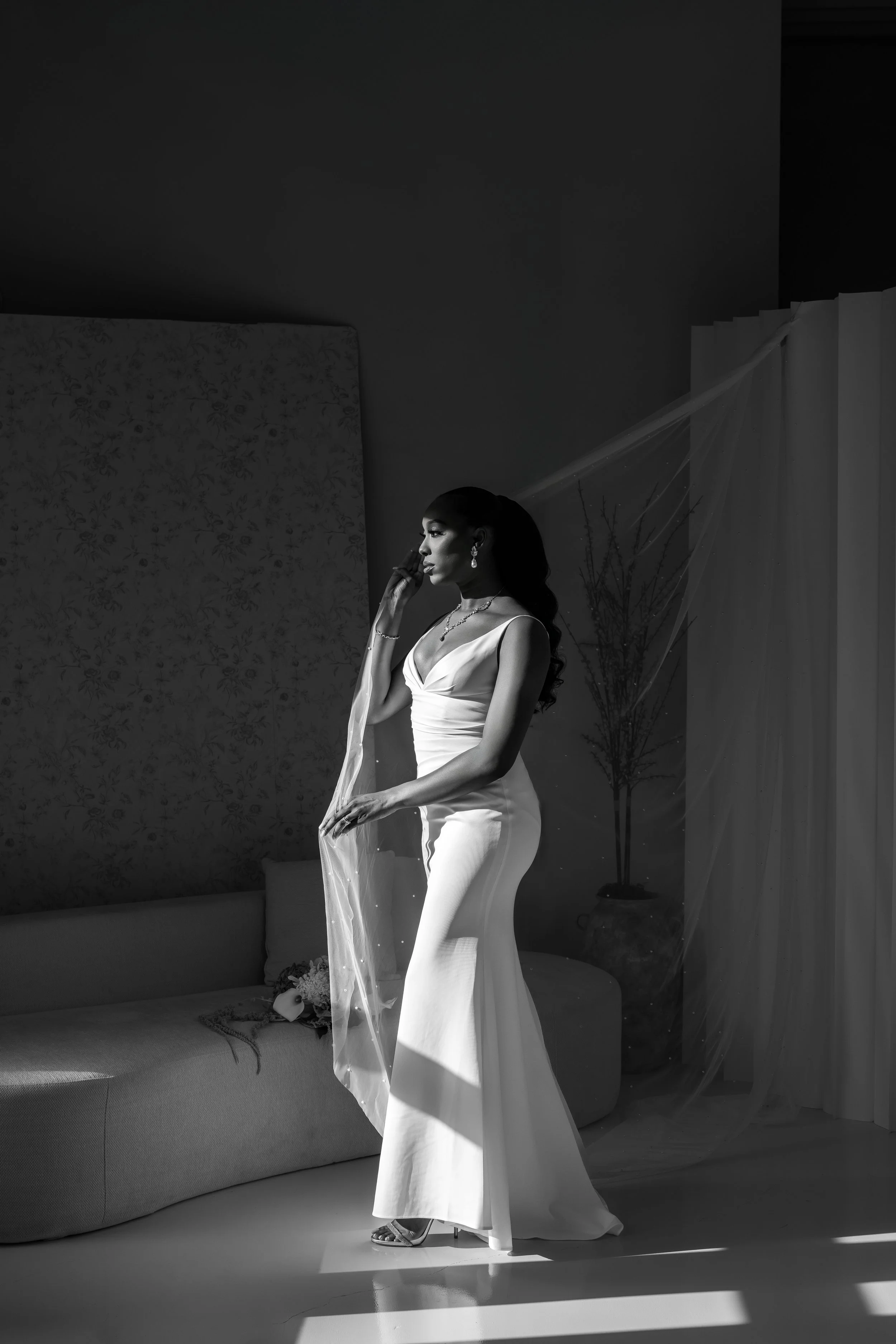 A woman in a white dress stands elegantly in a room with shadows and soft lighting, holding a veil and surrounded by minimal decor.