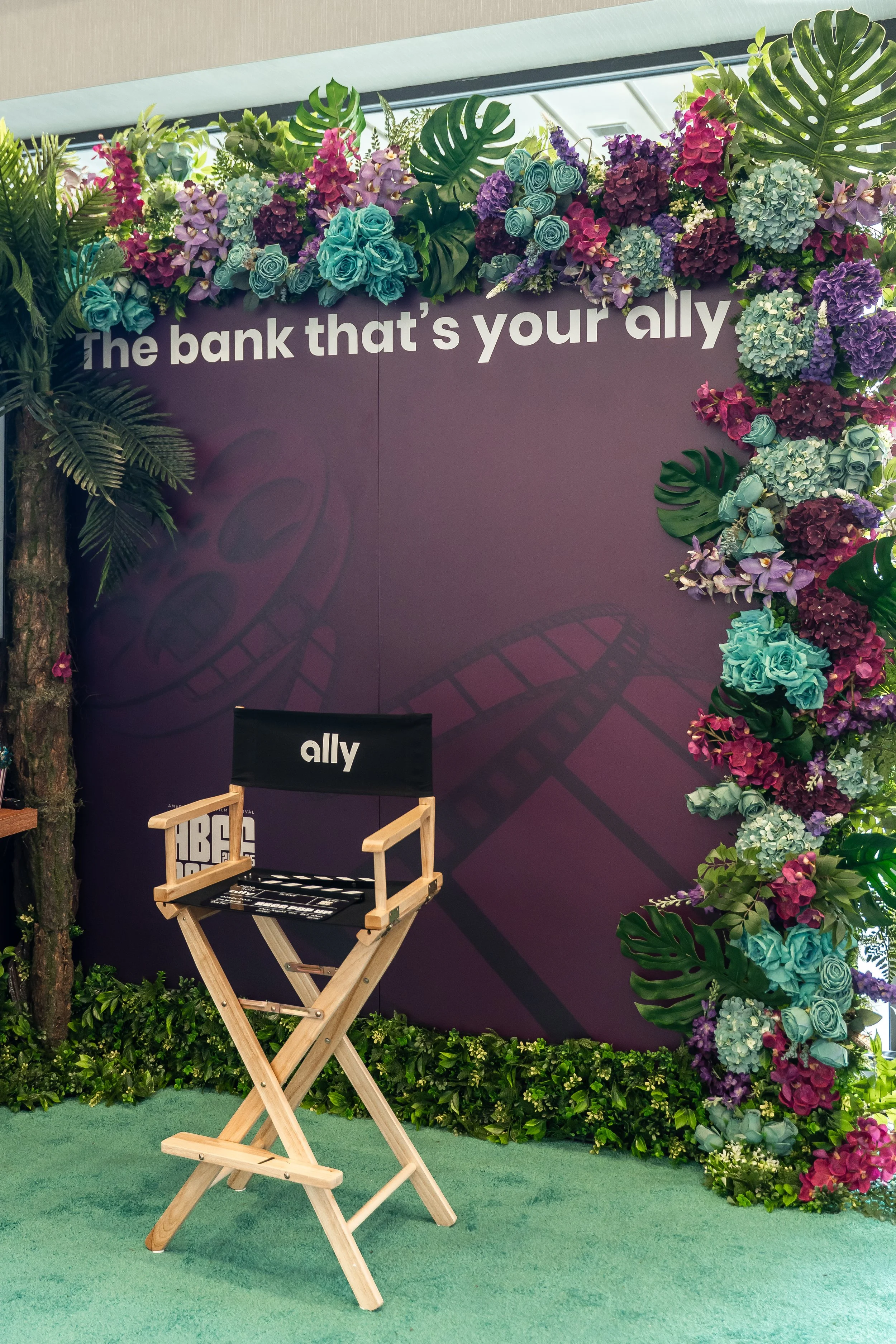 A decorated wall and stage setup with a purple background, floral arrangements of pink, purple, and blue flowers, and large green tropical leaves. The wall displays the phrase 'The bank that’s your ally.' A wooden director's chair with black fabric a