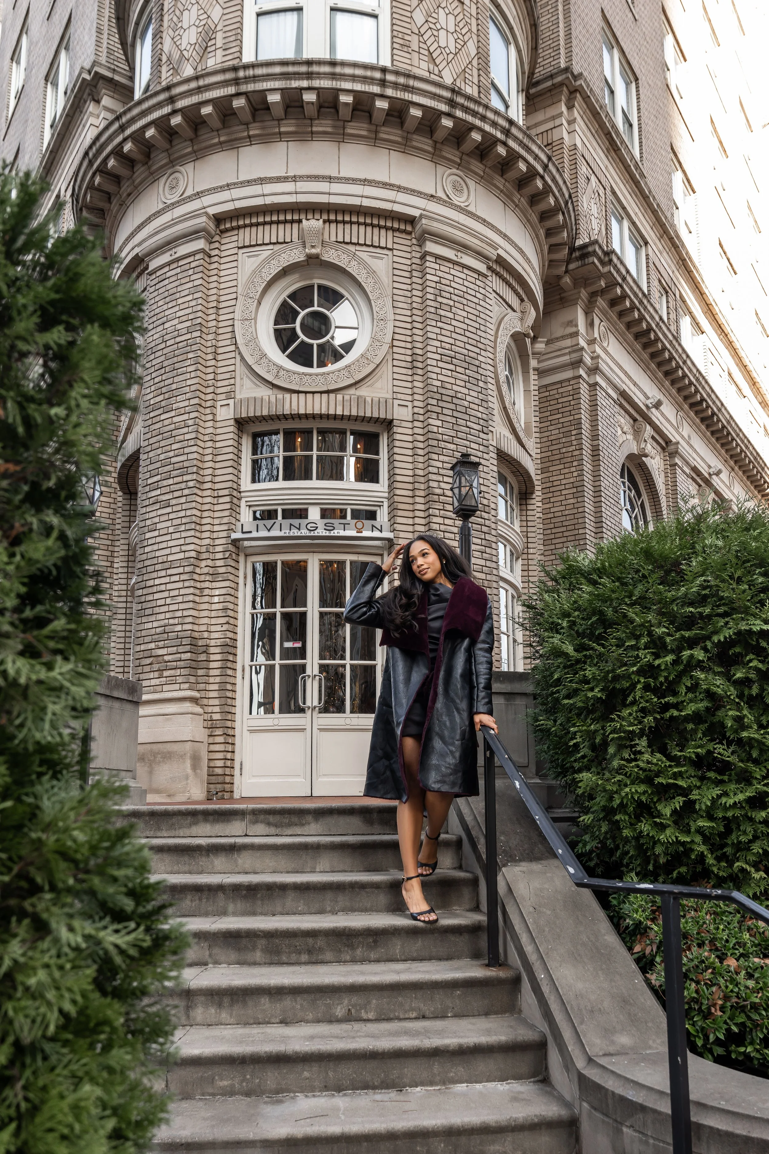 A woman dressed in a black dress, high heels, and a long black leather coat with purple fur accents, walking down stairs outside a large, ornate brick building with rounded windows and the sign 'LIVINGSTON' above the entrance.