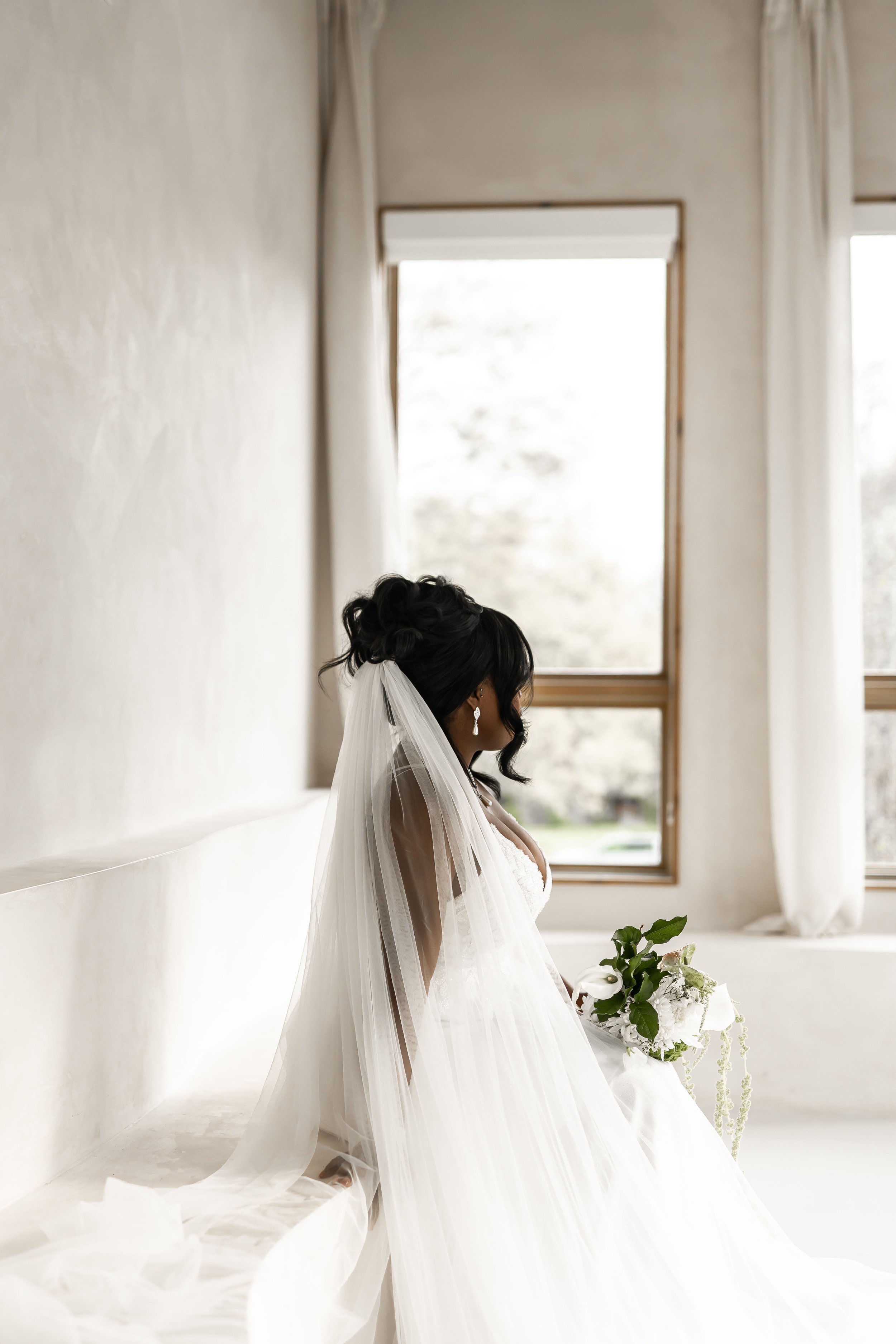 A bride sitting on a white bench, holding a bouquet, in a room with large windows and white curtains, with her head turned to the side.