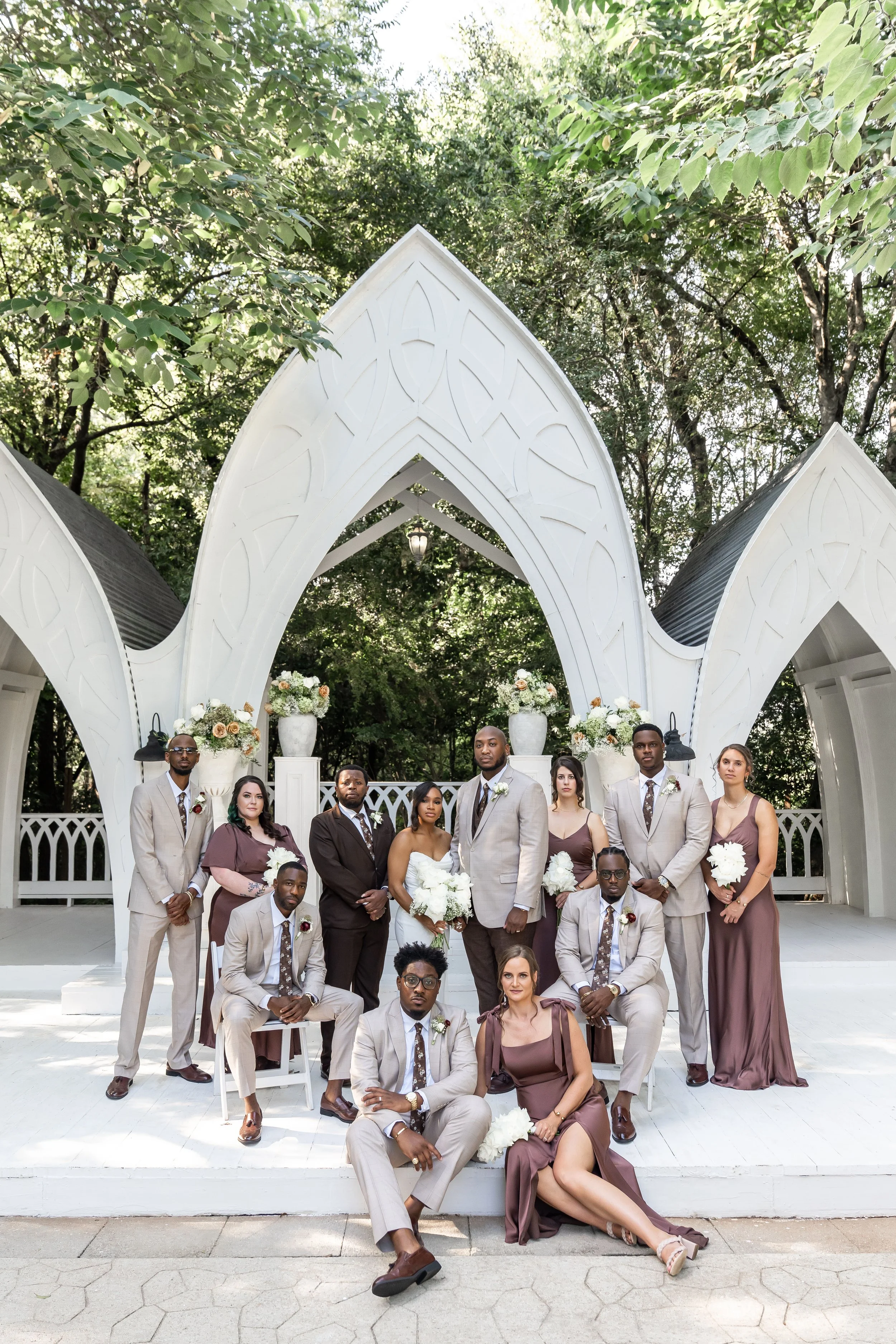 A wedding party with 13 people standing and sitting on a white platform in front of a Gothic-style white arch structure, with trees in the background. The bride is holding a bouquet, and the groom stands next to her. The group is dressed in formal at