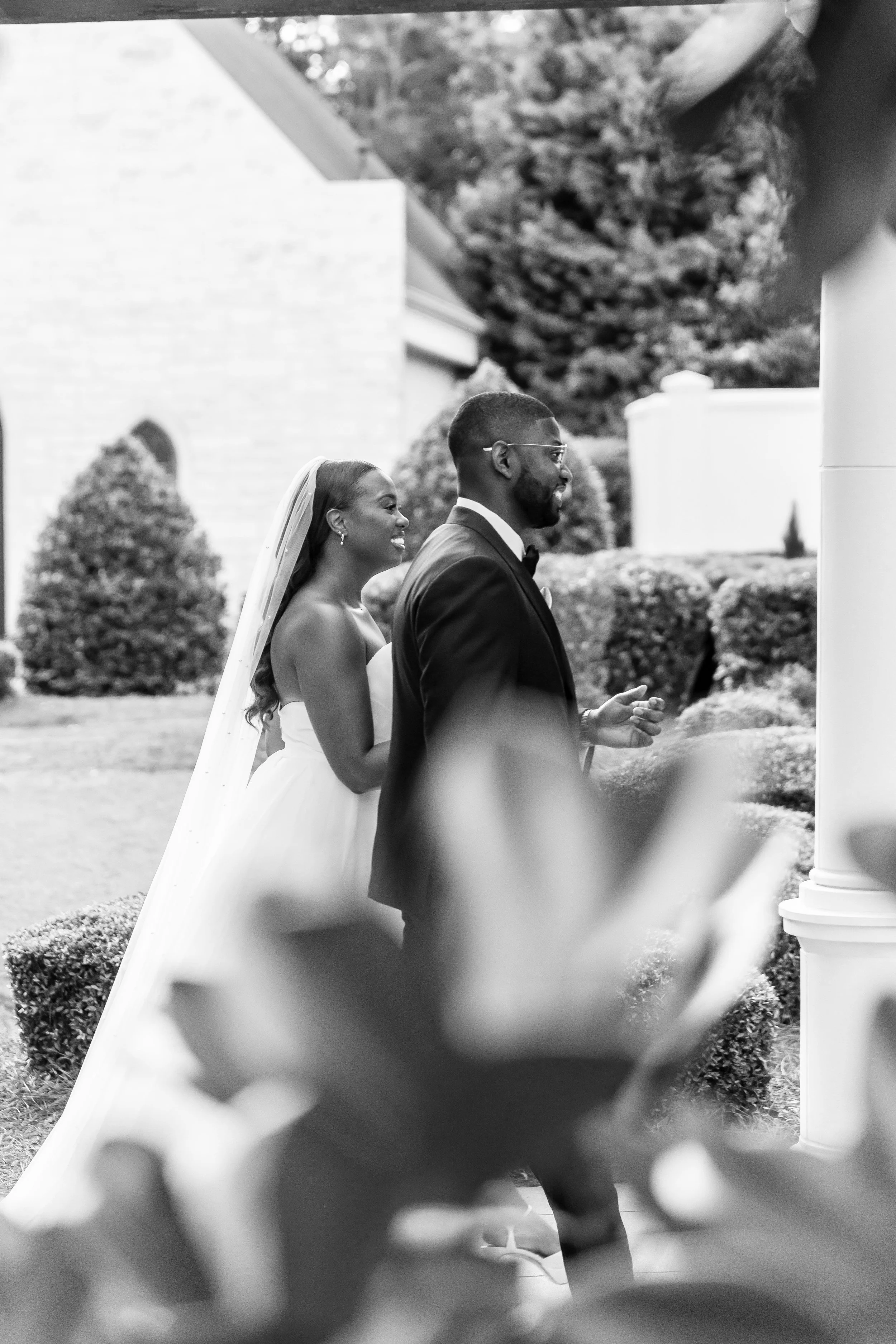 A black-and-white photo of a bride and groom smiling outdoors during a wedding ceremony, with the bride wearing a strapless wedding dress and veil, and the groom in a tuxedo with glasses, standing among hedges and trees.