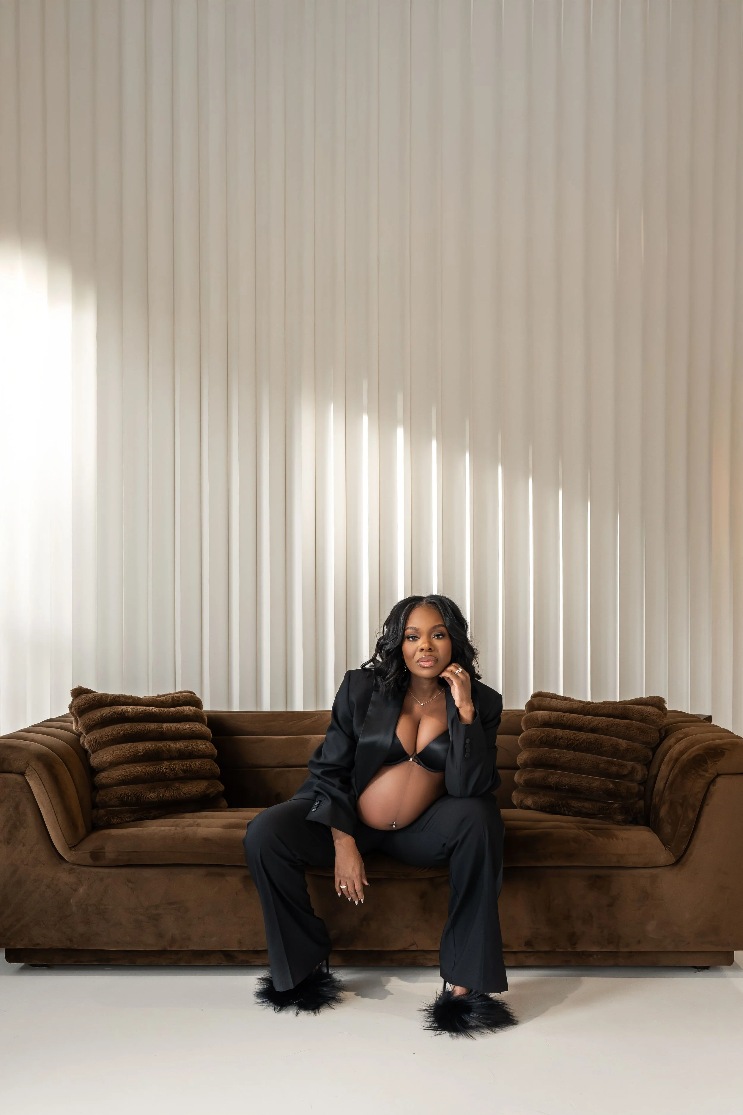 A pregnant woman in black lingerie and matching blazer and pants, sitting on a brown velvet sofa with brown pillows, in front of a white curtain backdrop.