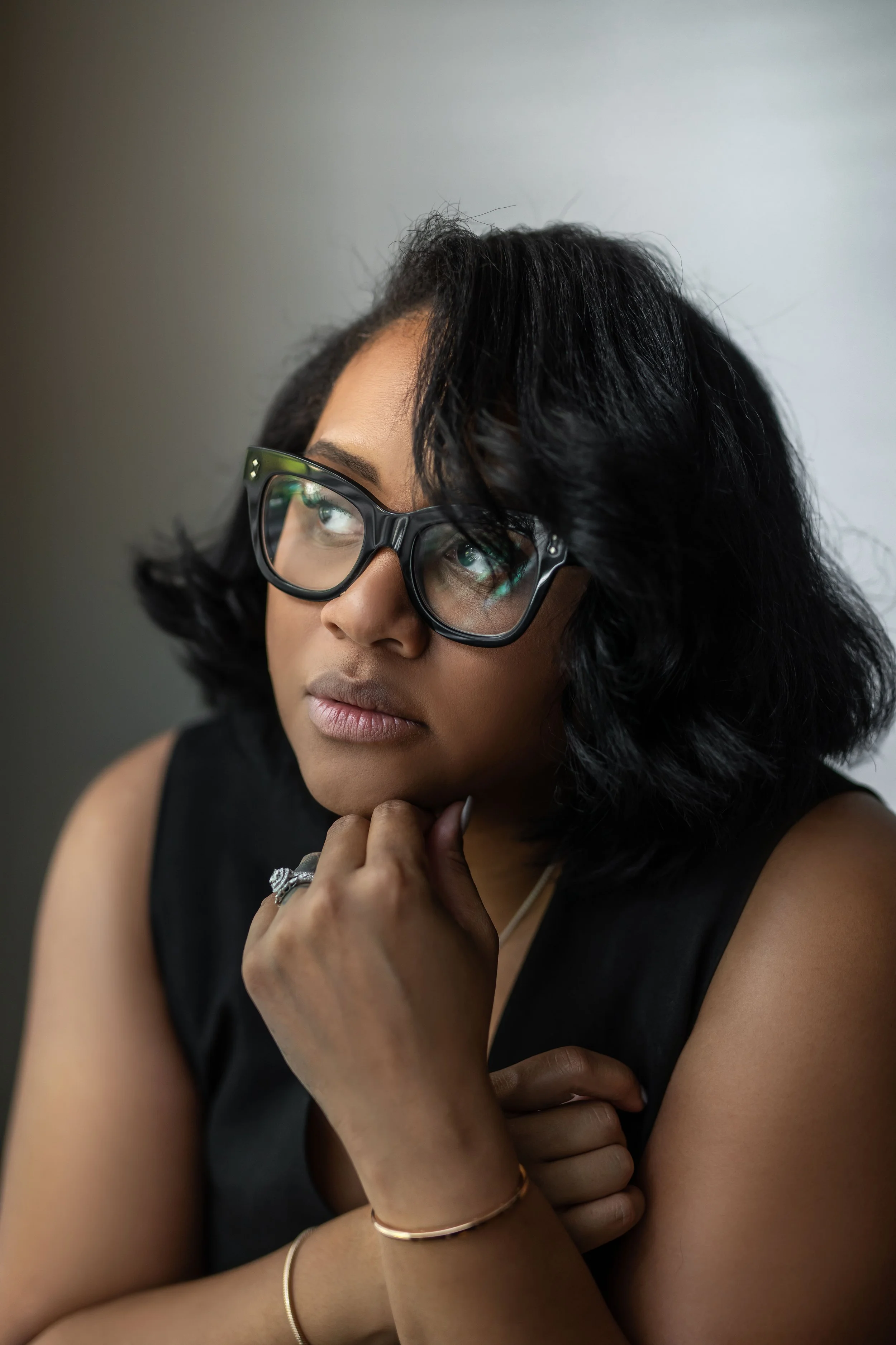 A woman with black, wavy hair and glasses, resting her chin on her hand, looking thoughtfully to the side.