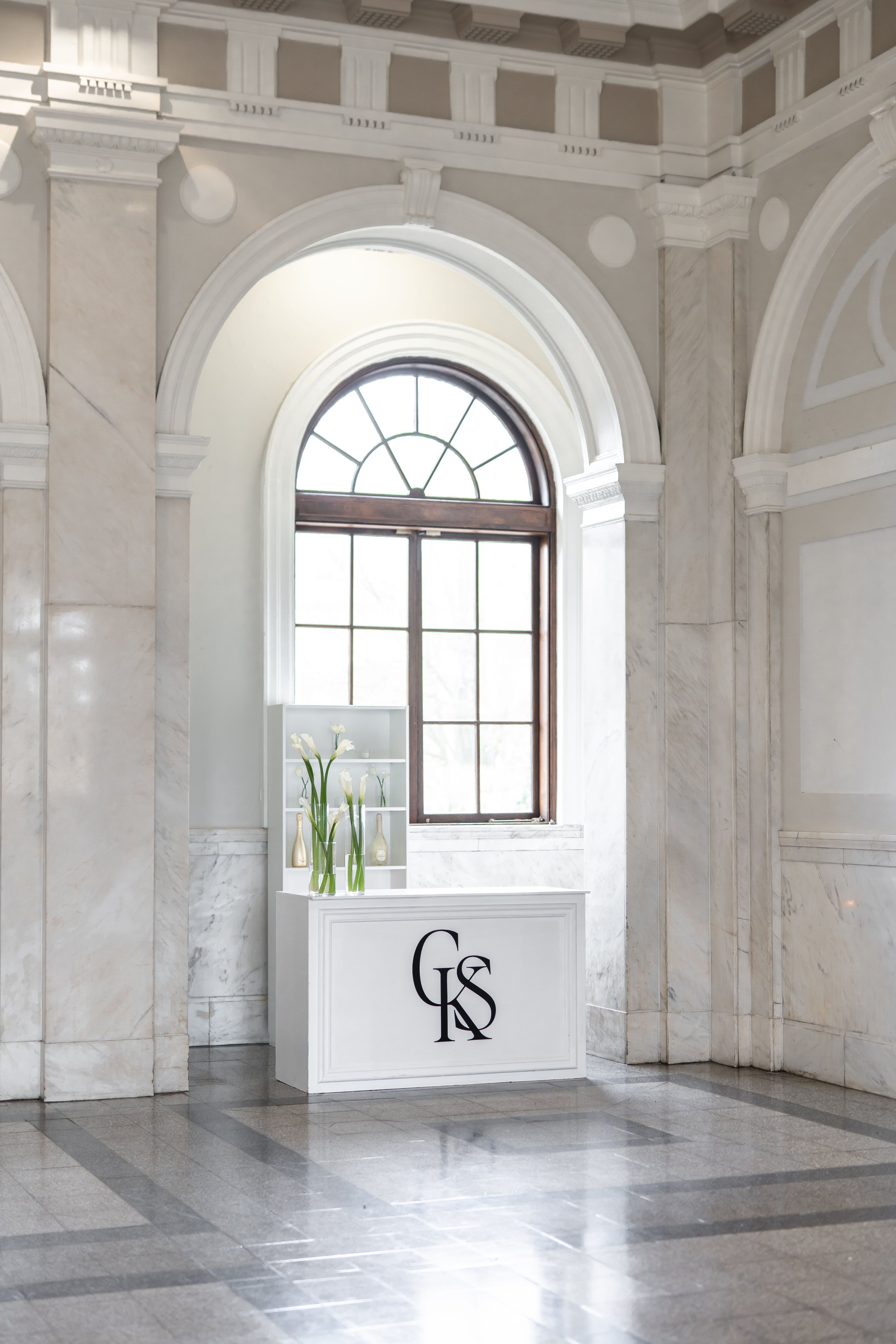 Interior of a building with marble walls, a large arched window, and a white display stand with vases and tall white flowers. The display stand has the initials 'GKS' in black.