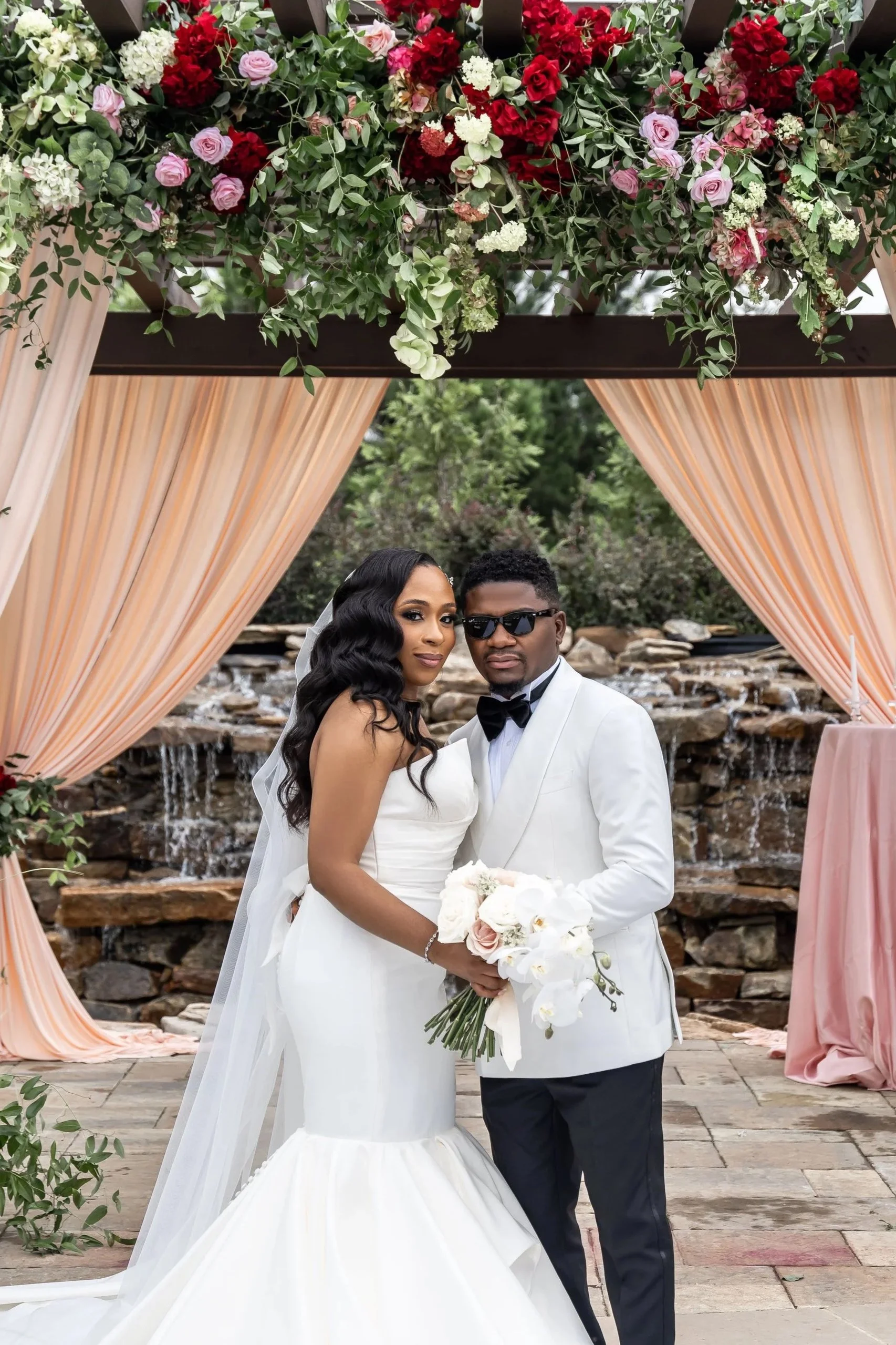 A newlywed couple in wedding attire, standing under a floral wedding arch with peach-colored drapery, outdoors near a waterfall feature.