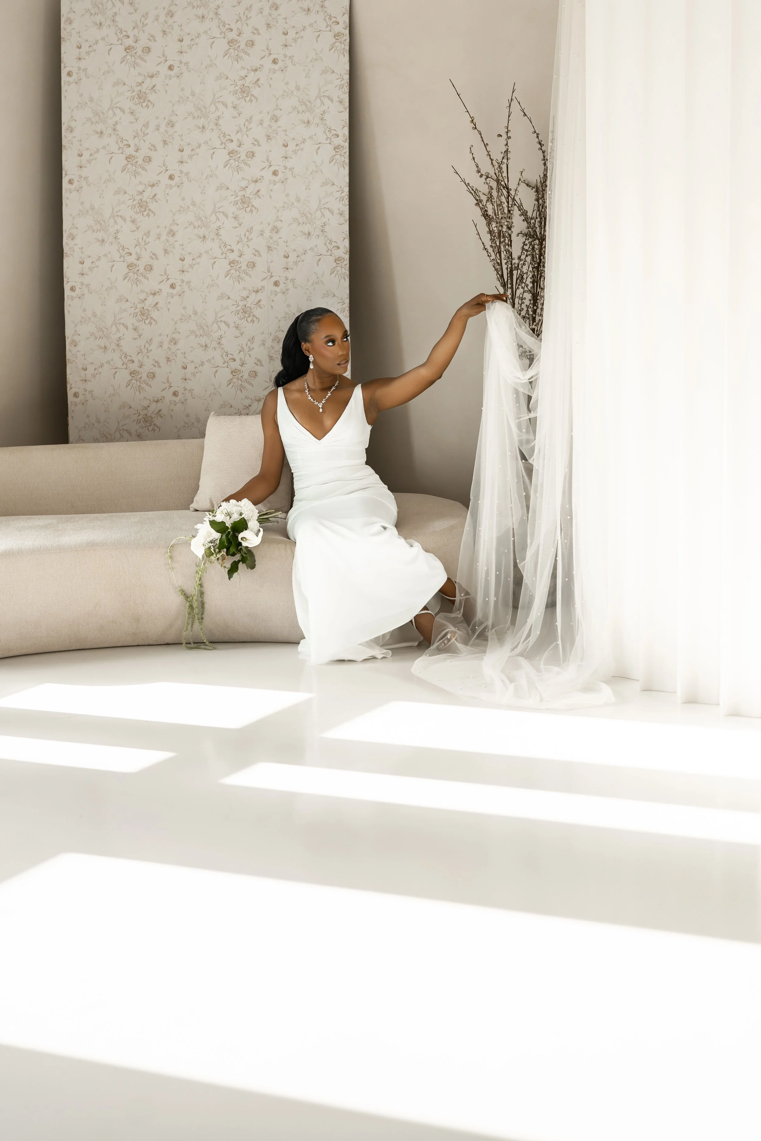 A bride in a white wedding dress sits on a beige sofa holding a bouquet of white flowers, with her right hand reaching towards a sheer white curtain.