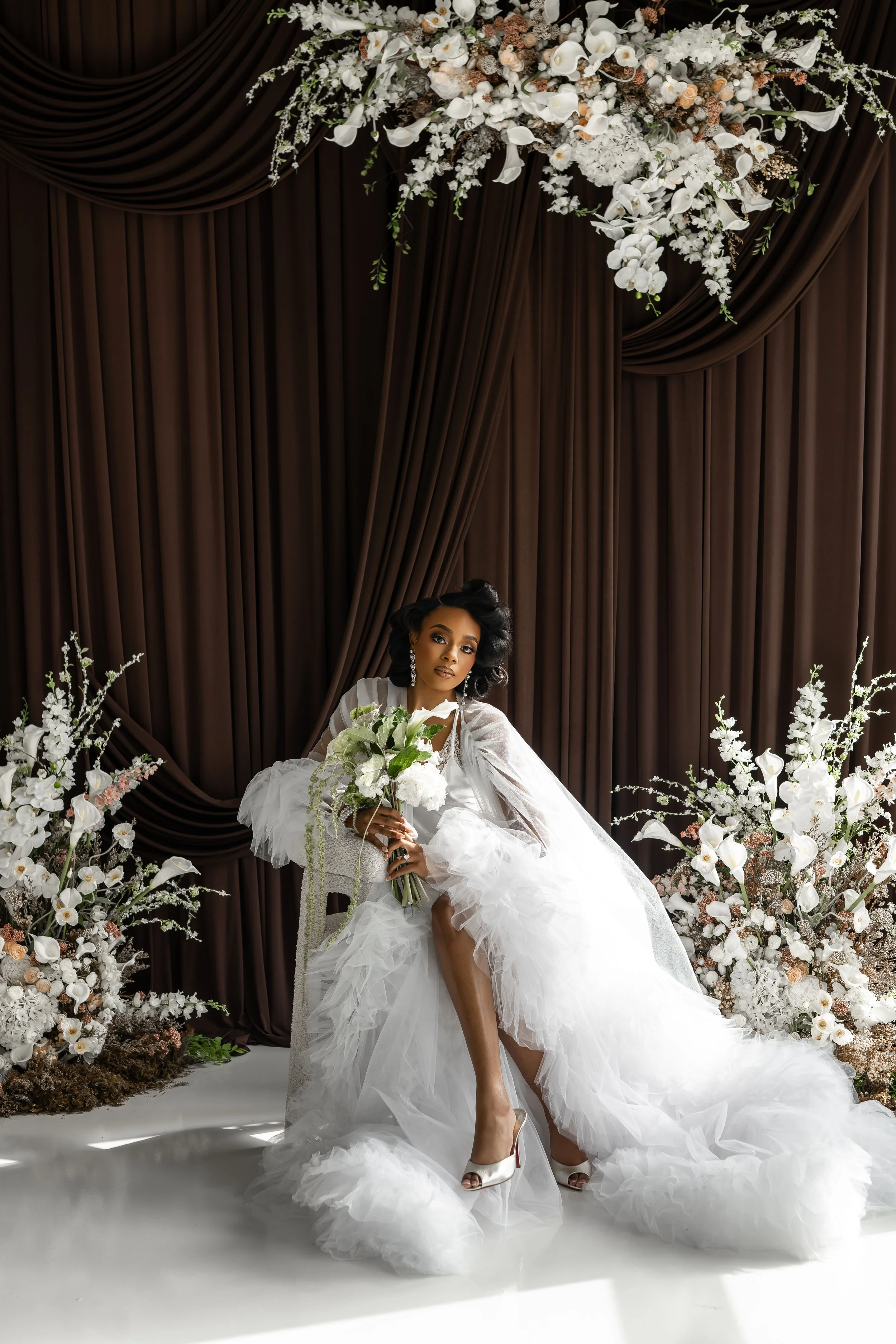 A woman in a white wedding dress with tulle details poses with a bouquet of white flowers in front of dark brown draped curtains and floral arrangements of white flowers and greenery.