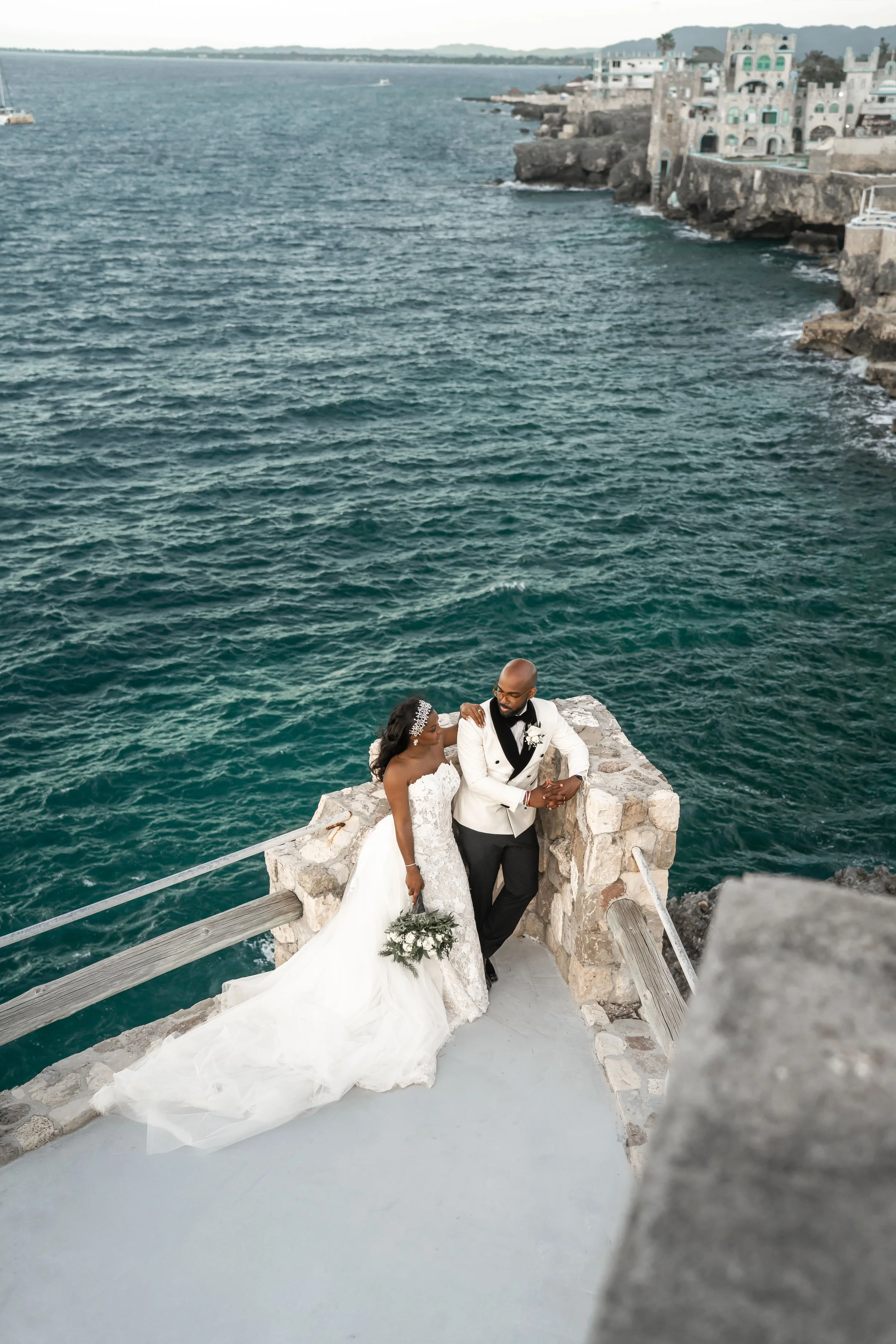 A bride and groom are standing by a stone railing on a seaside overlook, with the ocean and cliffs in the background. The bride is wearing a white strapless wedding gown and holding a bouquet, while the groom is dressed in a tuxedo with a black bow t