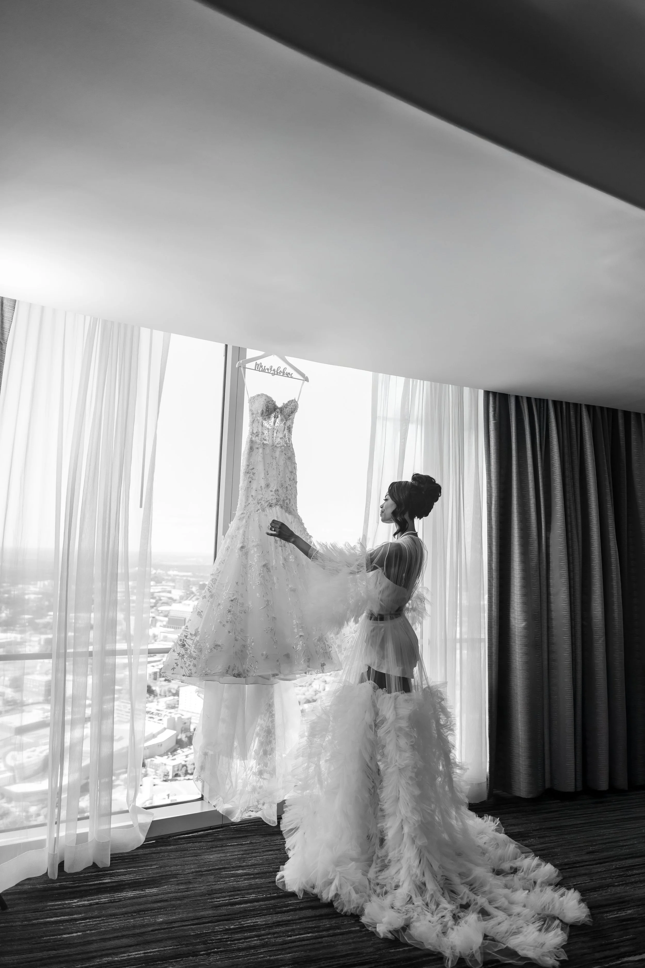 A bride dressed in a sheer, feathered gown looks at a wedding dress hanging in front of a large window with city views.