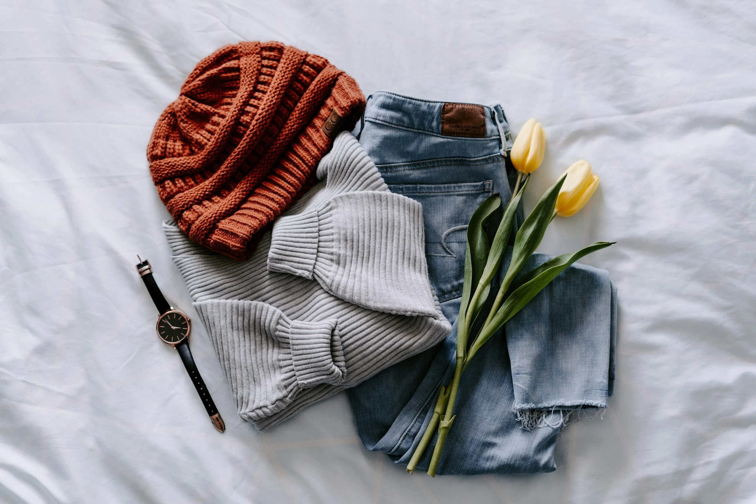 A flat lay of casual clothing and accessories: a rust-colored knit hat, a gray ribbed sweater, a pair of blue jeans, a black watch with a gold face, and three yellow tulips with green stems and leaves, all arranged on a white surface.