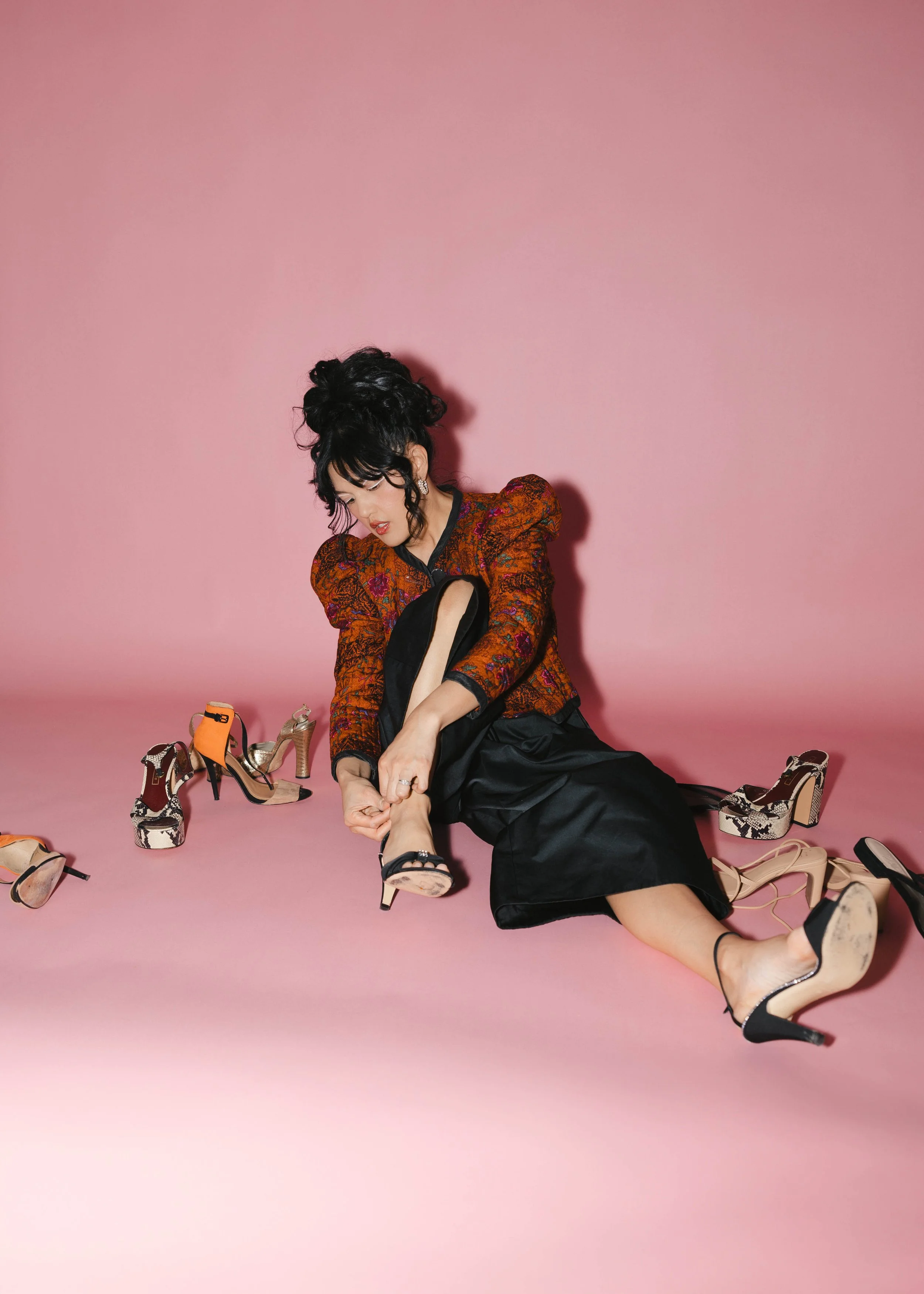 Woman sitting on pink floor with multiple high-heeled shoes, adjusting one shoe on her foot, wearing a patterned orange and black jacket and black skirt, against a pink background.