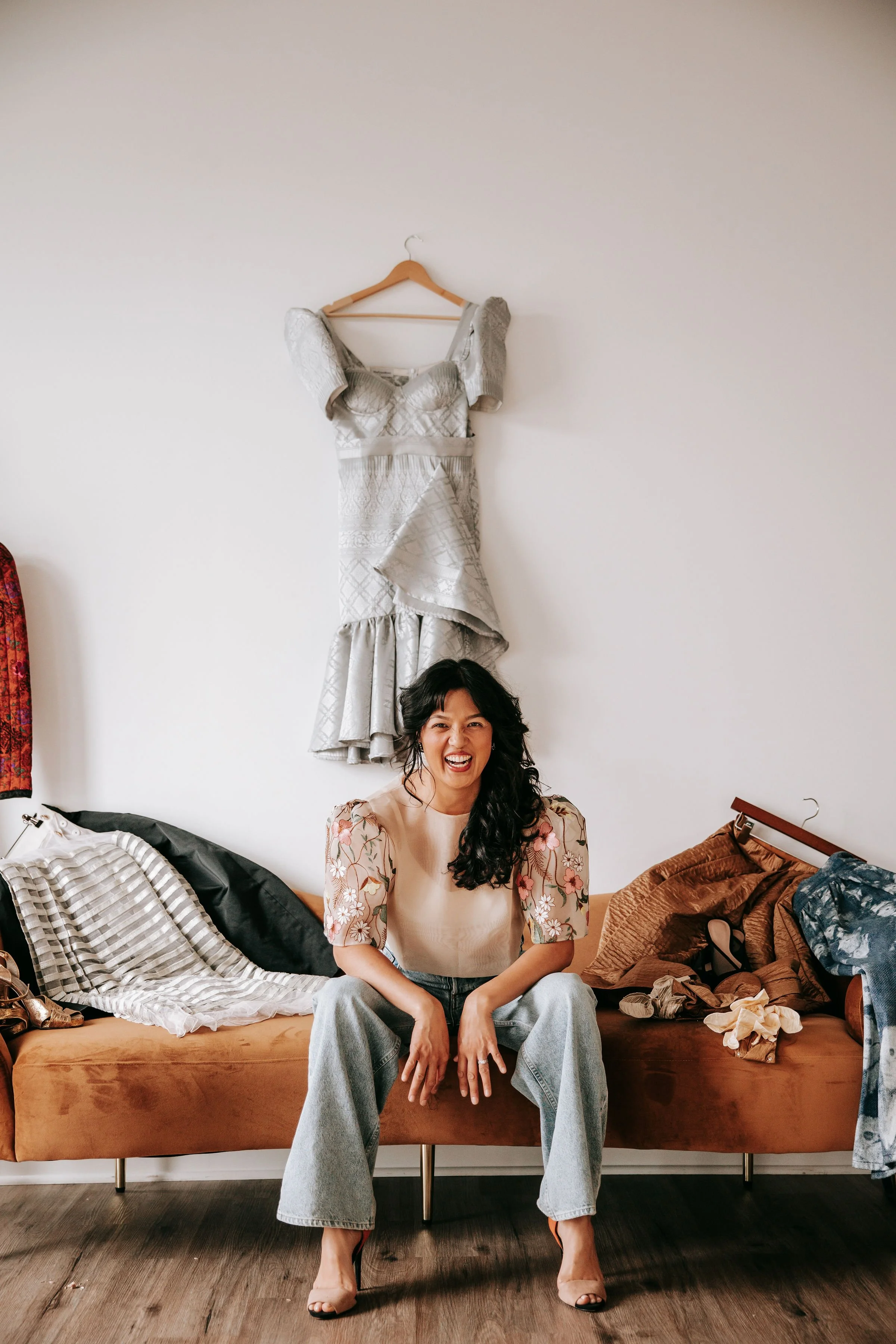 Woman sitting on a brown sofa with messy clothes and shoes around her, smiling and showing her teeth.
