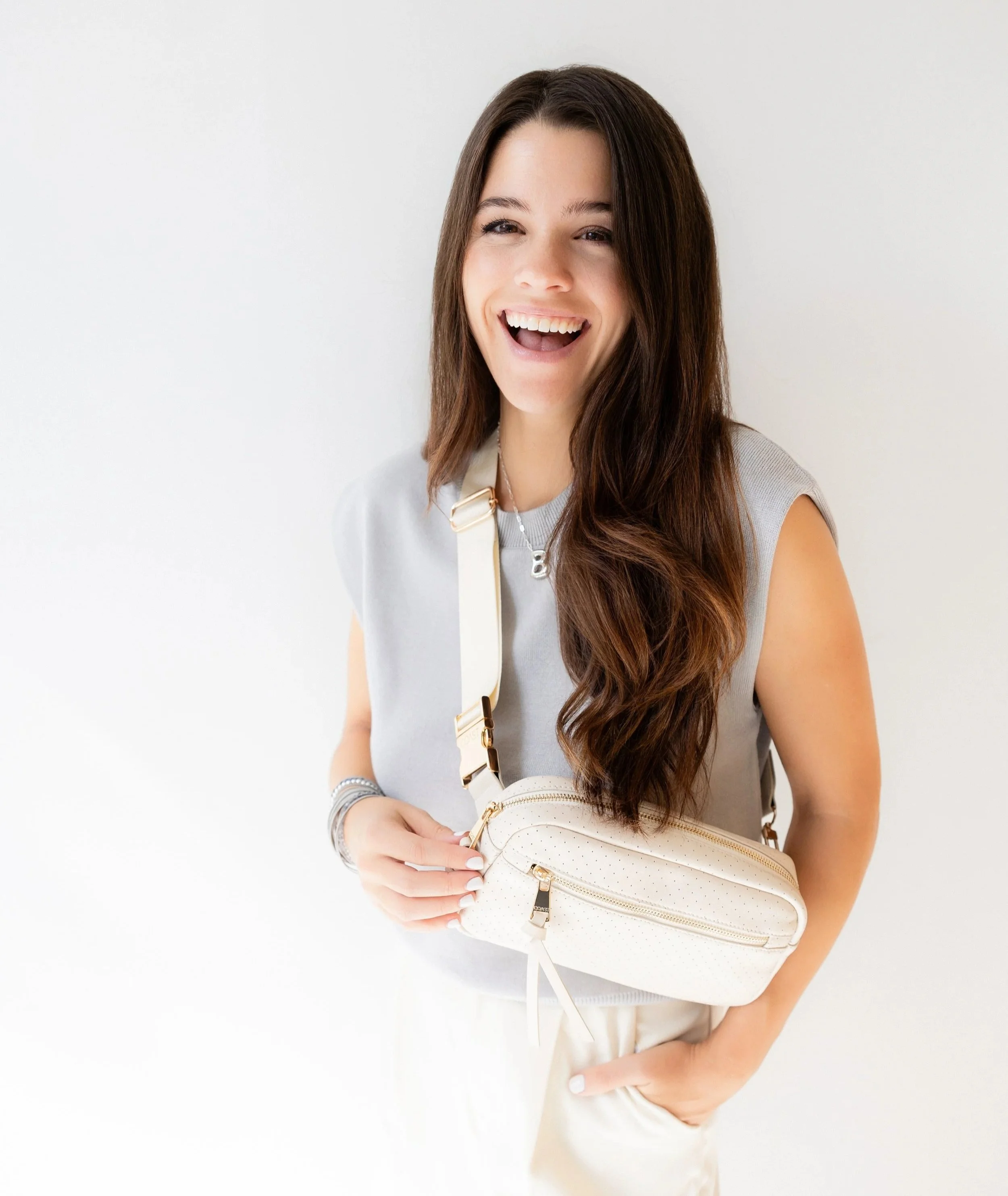 A smiling young woman with long brown hair, wearing a sleeveless gray top, holding a white purse, and standing against a plain white background.