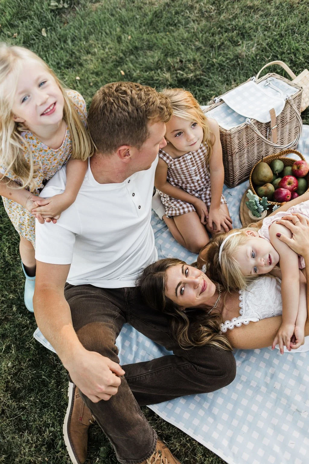 A family of six having a picnic outdoors on a grassy field, sitting on a blue checkered blanket with a basket of apples and a container of watermelon, with one kid hugging her mom.