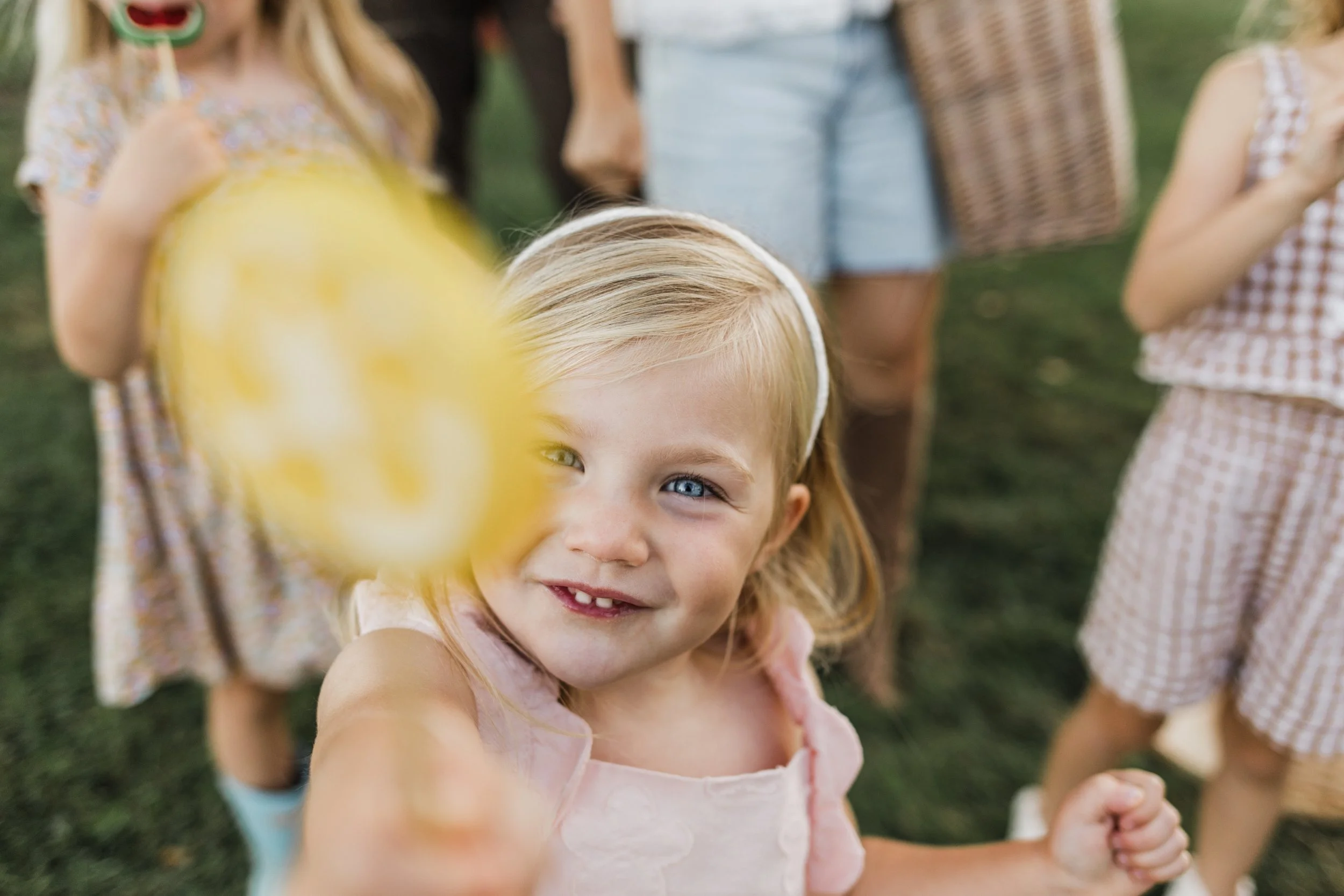 A young girl with blonde hair and blue eyes holding a yellow balloon with a smiling face printed on it, at an outdoor gathering with other children in the background.