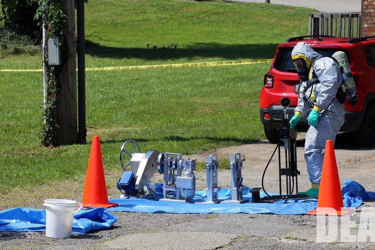 DEA hazmat team member in a protective suit next to seized pill press equipment outside a clandestine lab