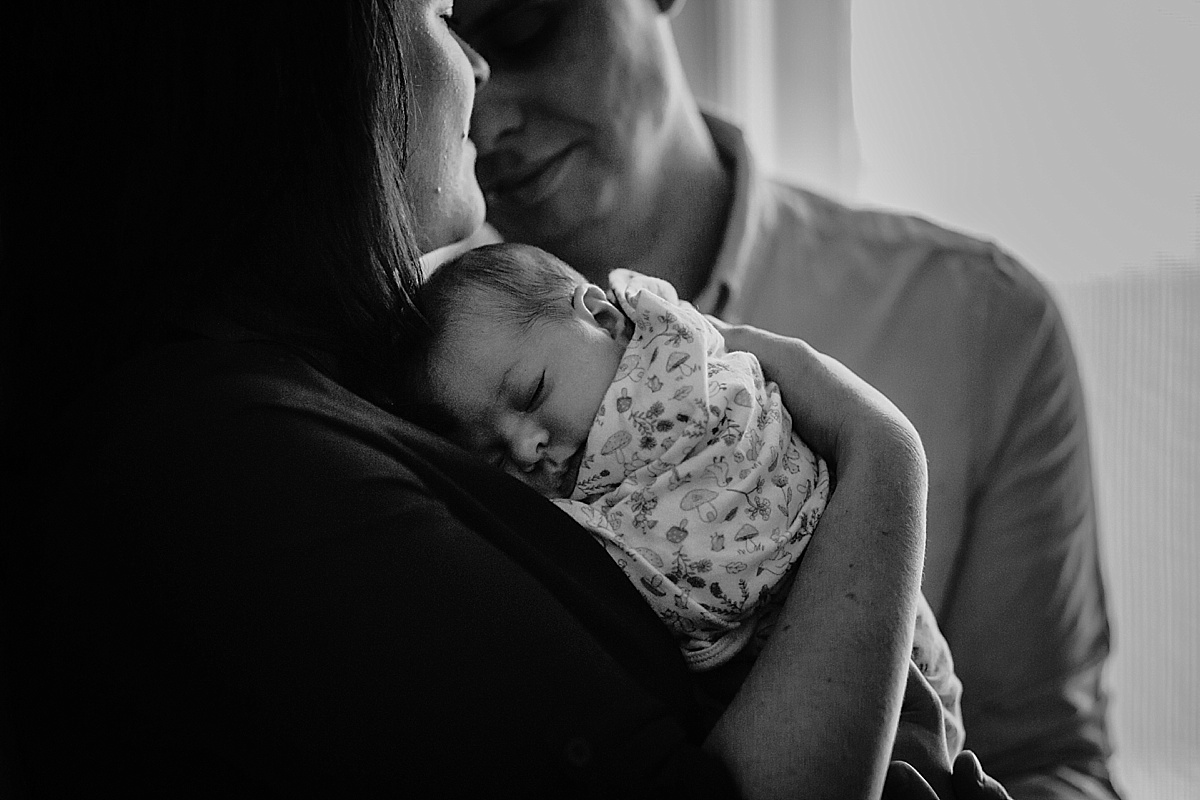 black and white image of baby laying on mums chest and dad leaning in