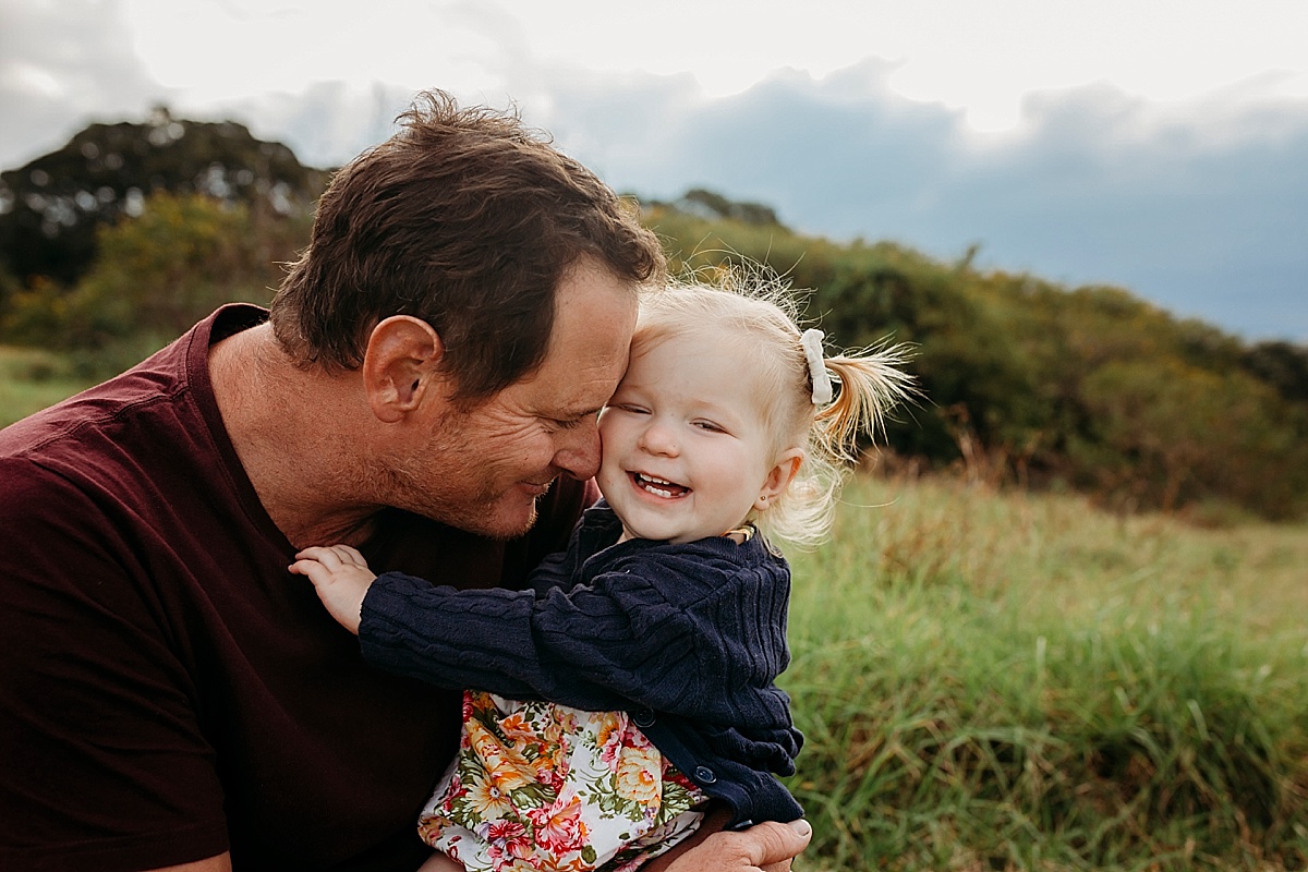 dad resting head against baby girl with baby girl looking at camera laughing