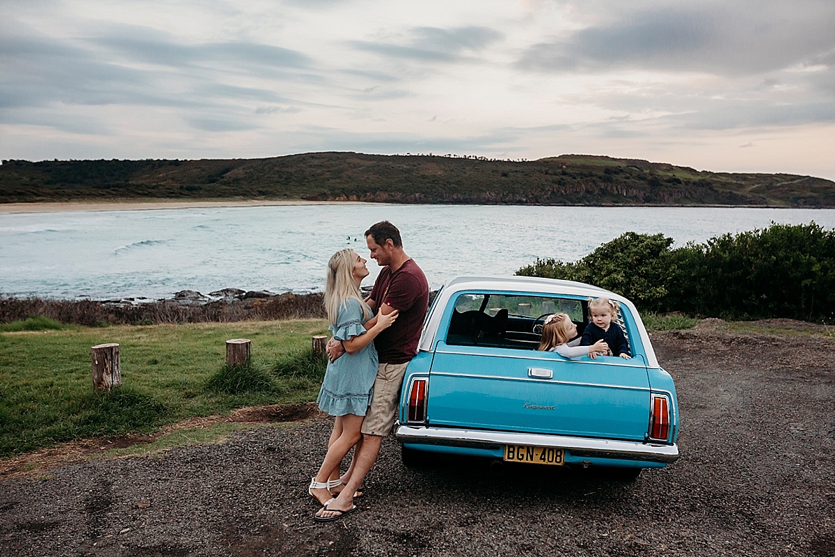 Mum and dad leaning against car hugging, daughters in back of car leaning on tailgate