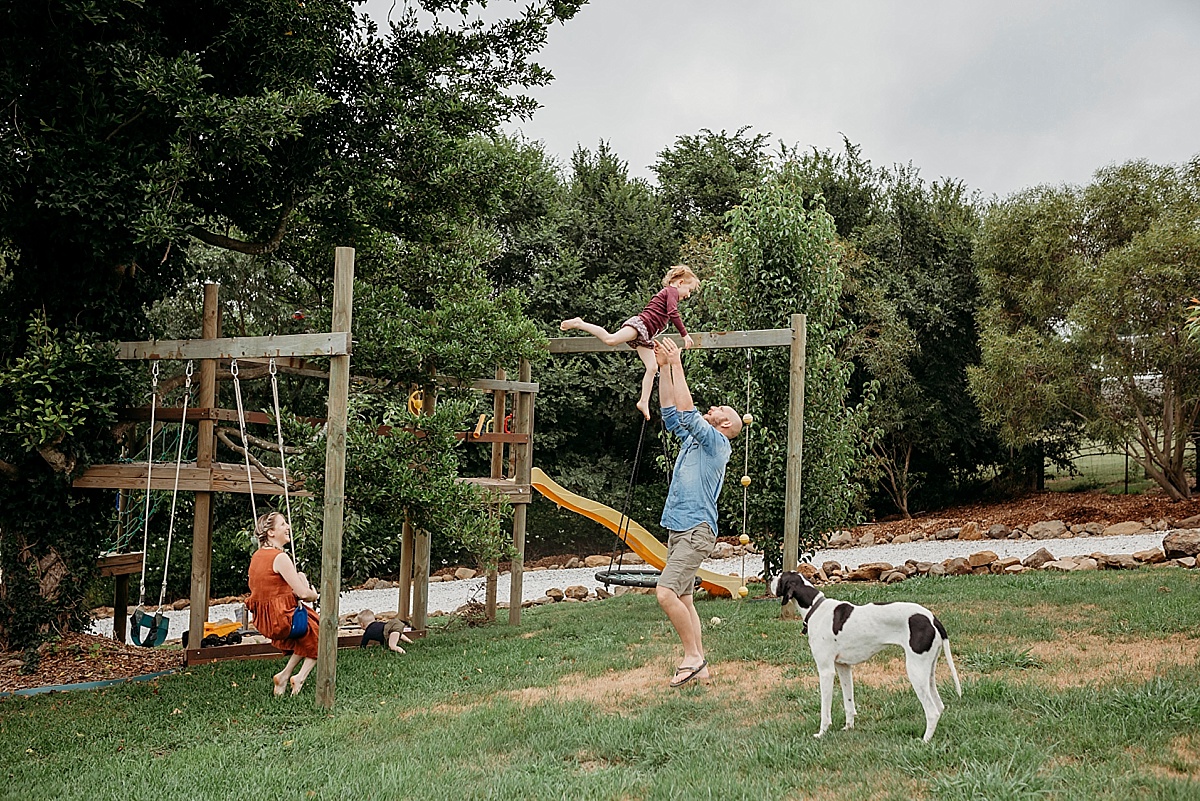 family in front yard, mum holding newborn sitting on a swing, son in sandpit, dog laying on the ground, father throwing daughter in the air