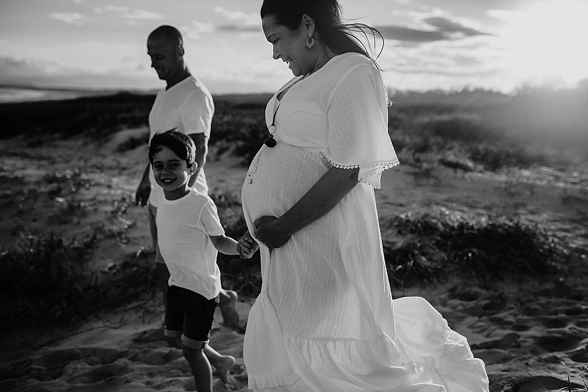 black and white image of family of 3 holding hands walking down the beach, pregnant baby. 