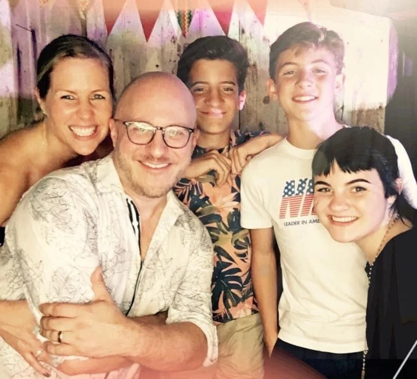 Group of five smiling people, including three teenagers and two adults, at a social gathering. They are indoors with a decorated background.